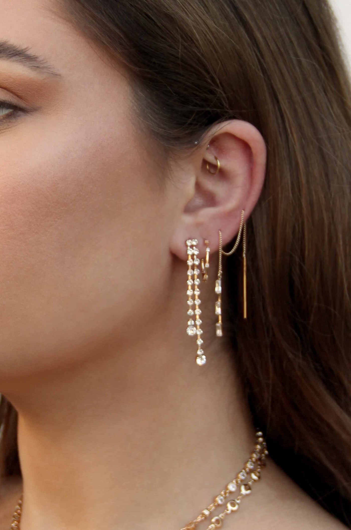 A close-up view of a woman's ear adorned with multiple earrings. Sparkling dangling earrings hang from her lobe, while delicate gold hoop and chain earrings add elegance, set against her smooth, light-toned skin.
