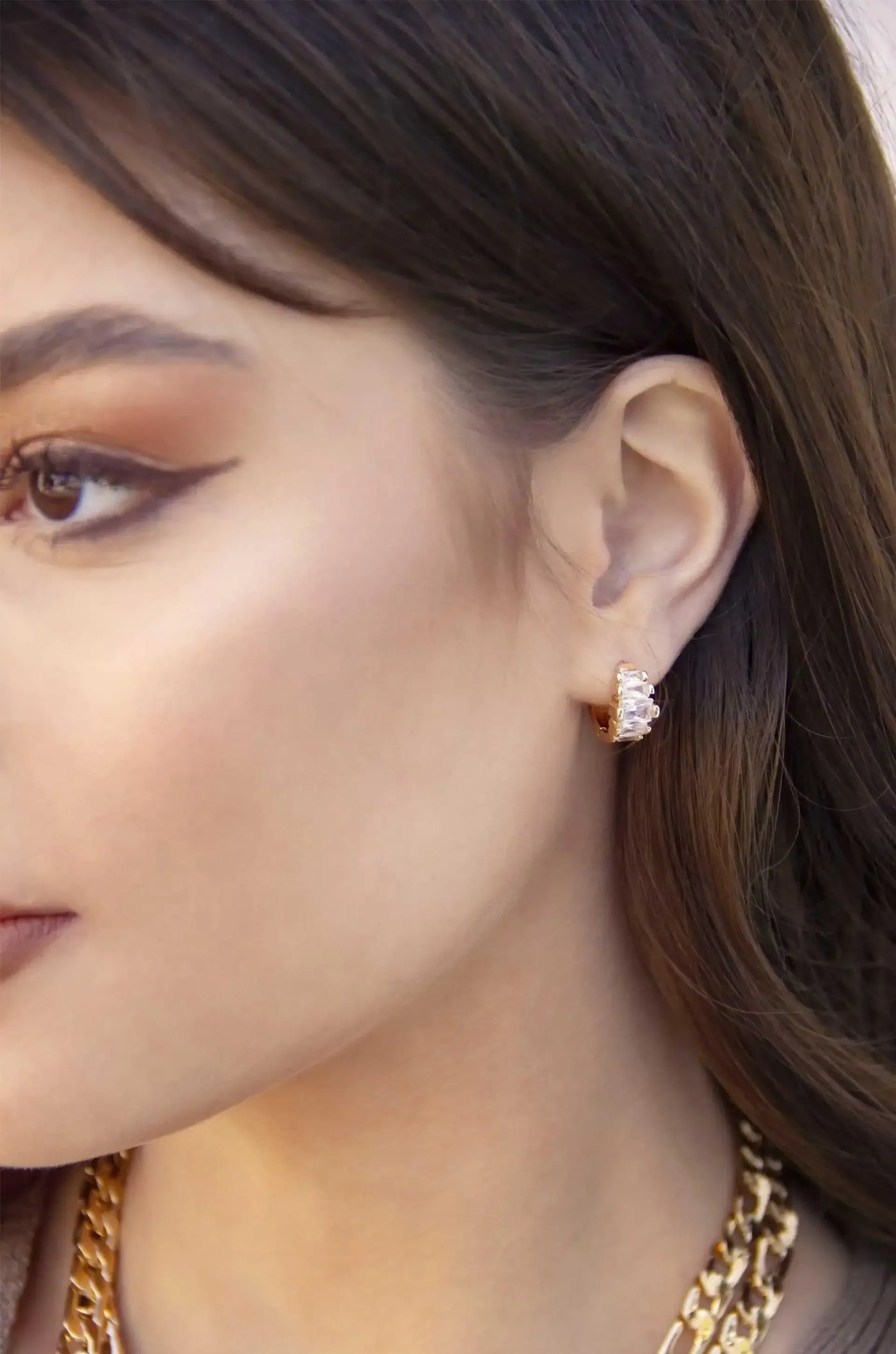 A close-up of a woman's profile highlights a crystal-adorned earring on her ear. Her skin has a soft glow, and she wears a chunky gold chain necklace, set against a neutral background.