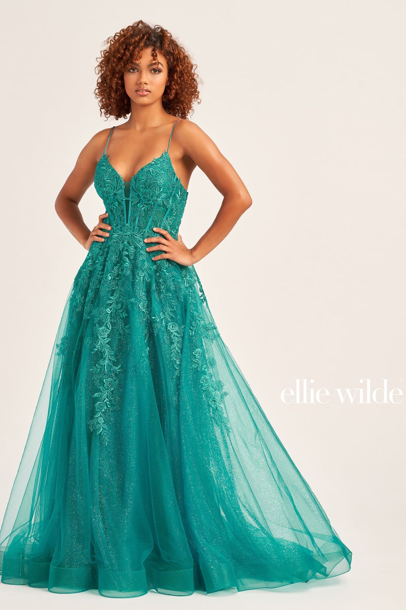 A woman modeling a teal green A-line evening gown with floral lace applique and spaghetti straps, photographed against a solid light beige background. The brand name 'ellie wilde' is visible in the bottom right corner.