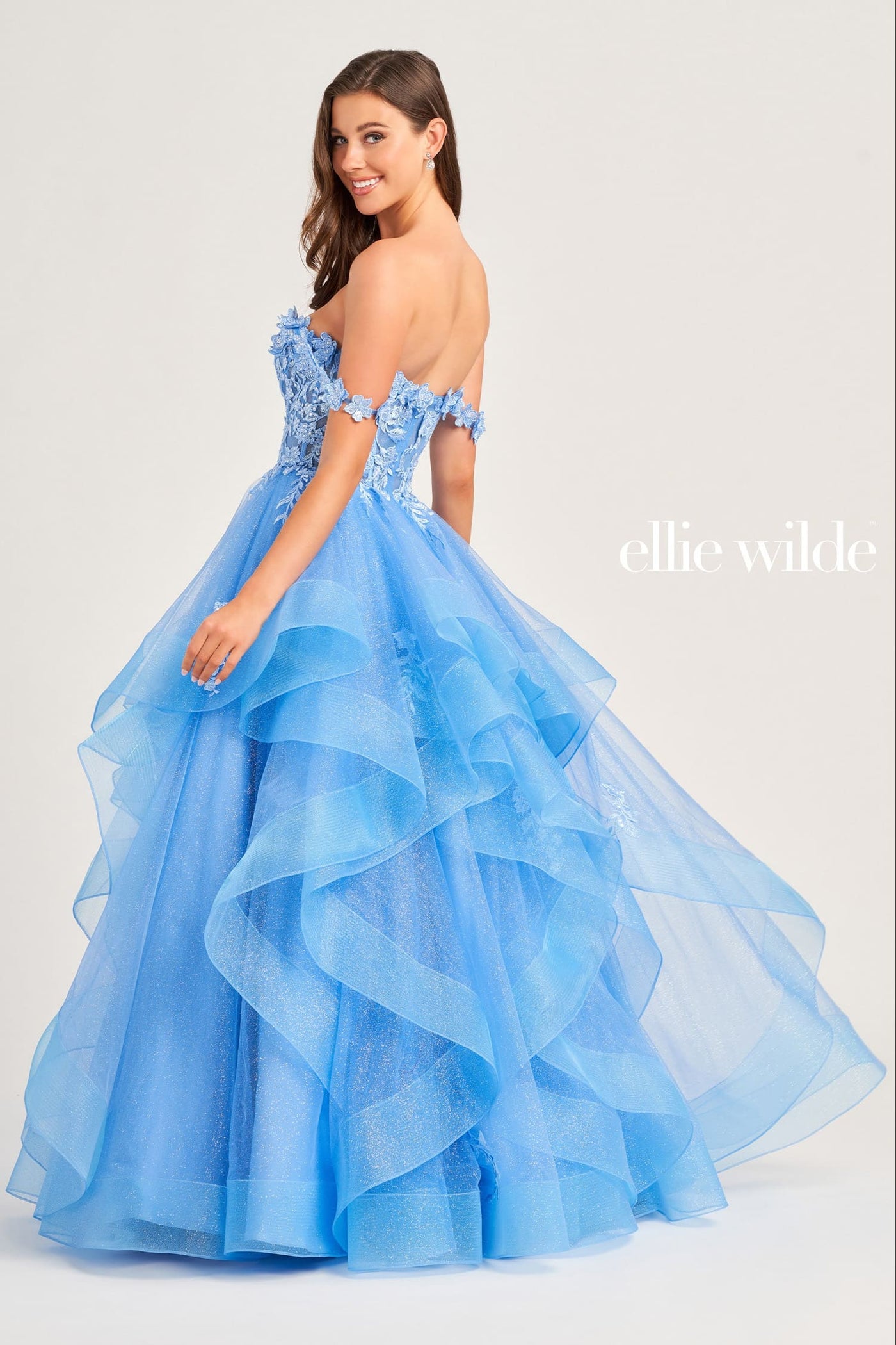A woman modeling an off-the-shoulder light blue ball gown with a layered ruffled tulle skirt, photographed against a plain light gray studio background. The brand name 'ellie wilde' appears in white text on the right side.