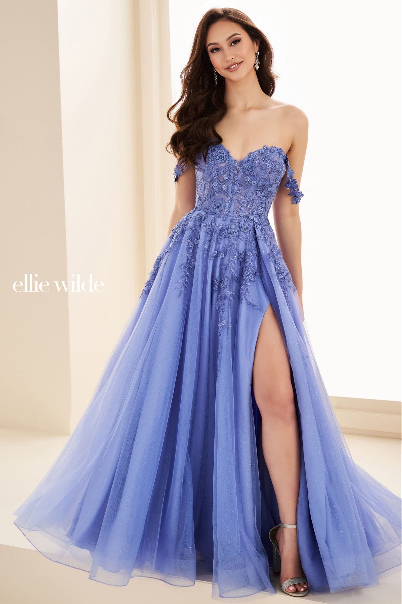 A woman models an Ellie Wilde strapless formal gown in a soft periwinkle blue. The gown features a sweetheart neckline, a heavily embellished lace bodice, off-the-shoulder lace sleeves, a high thigh slit, and a voluminous tulle skirt.