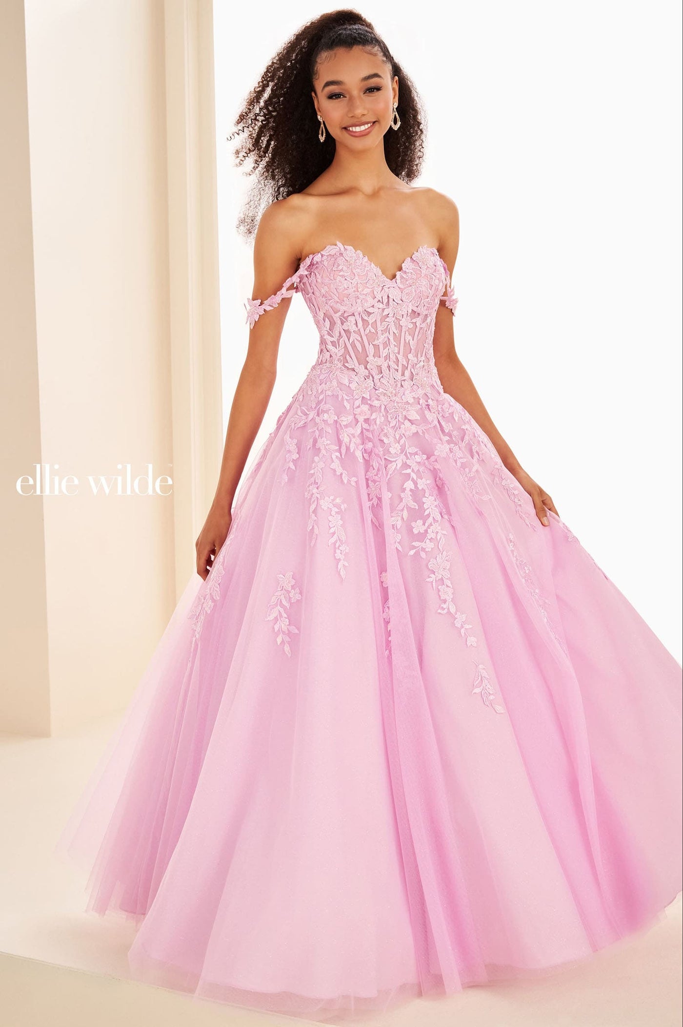 A woman modeling an off-the-shoulder pink ball gown with a fitted lace bodice and a full tulle skirt, photographed in a bright indoor setting. The brand name 'ellie wilde' appears in white text on the left side.