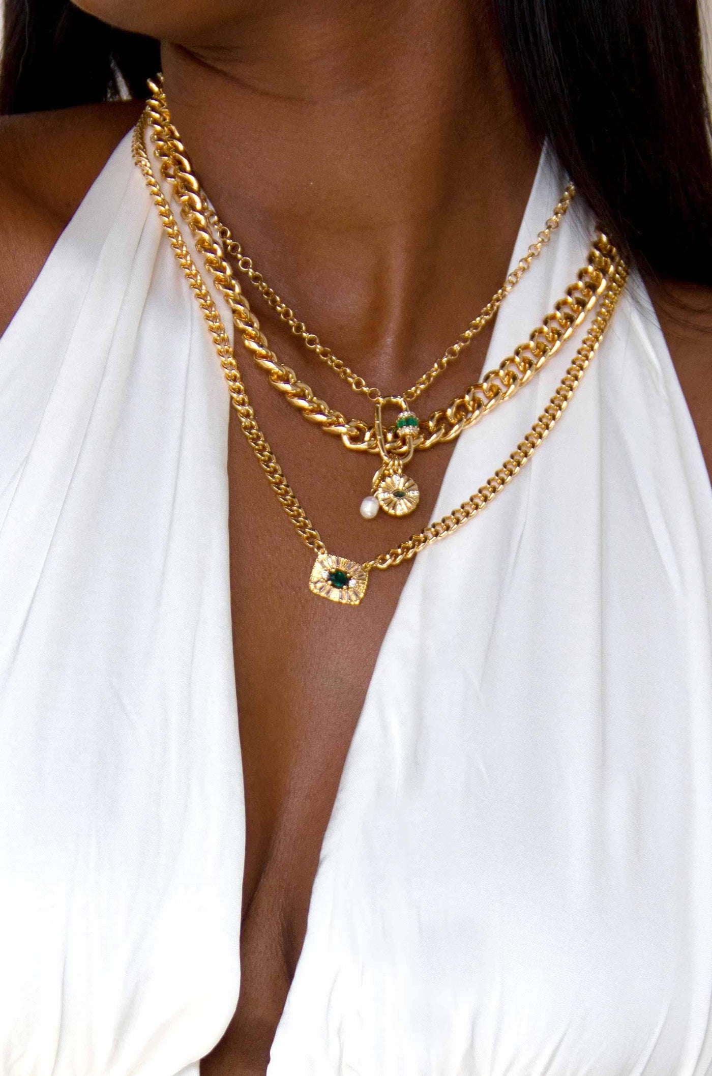 Three gold necklaces adorned with decorative pendants drape over tanned skin, accentuated by a deep V-neck white garment, highlighting the jewelry's intricate designs and gleaming finish.