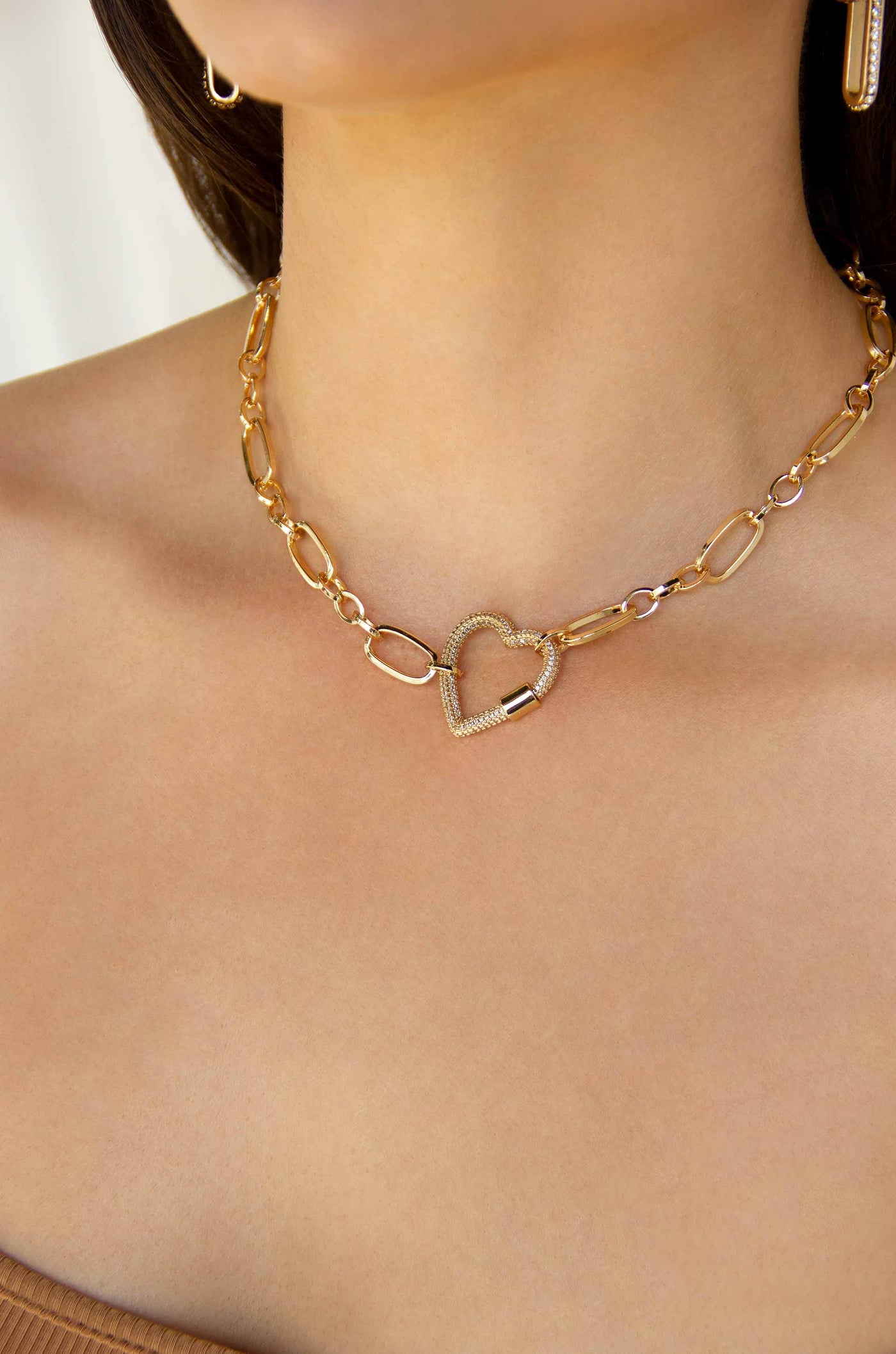 A gold chain necklace with a heart-shaped pendant rests on a bare shoulder, highlighting a smooth skin tone. The background features soft, neutral colors, enhancing the jewelry's elegance.