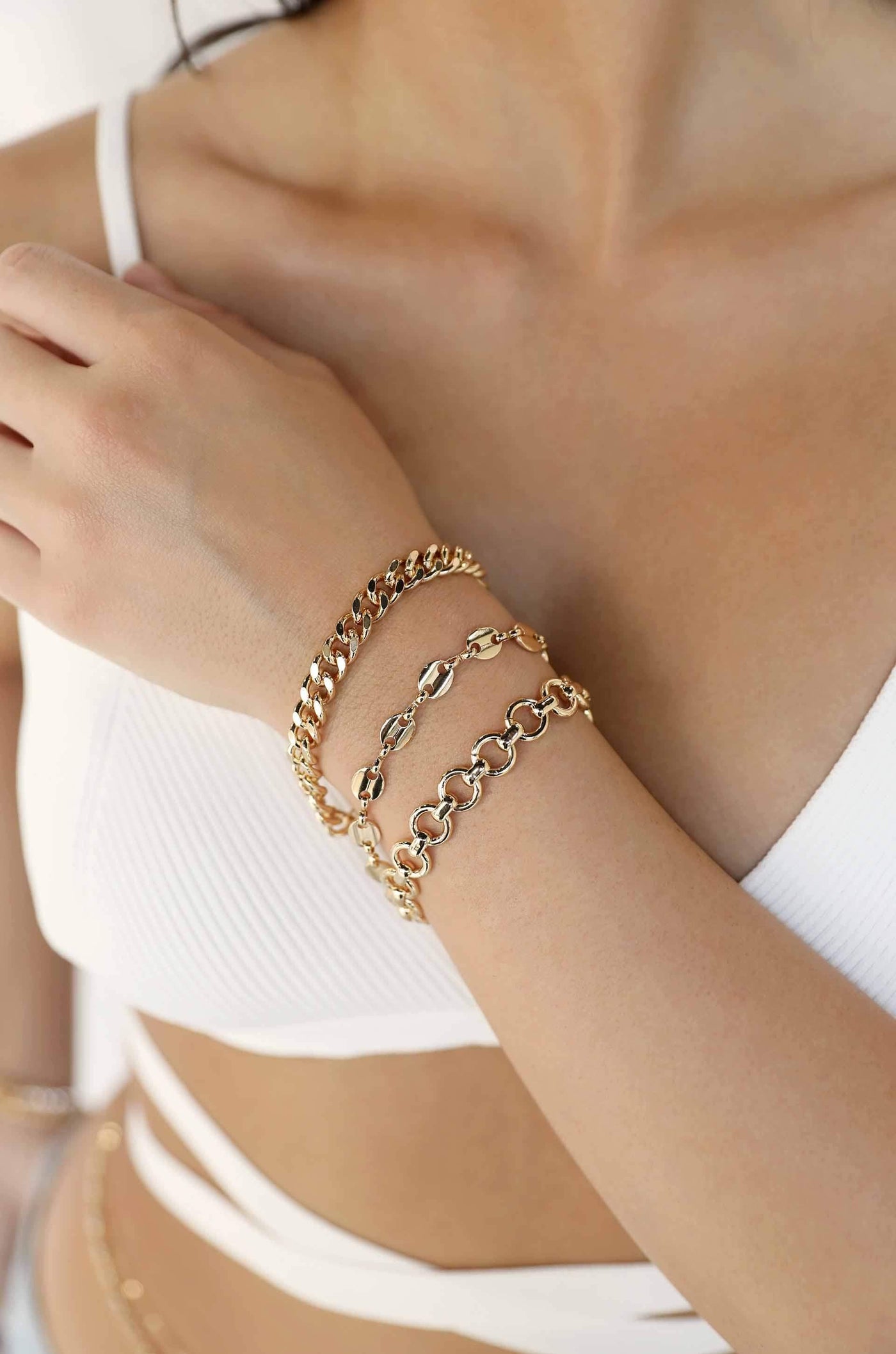 A hand adorned with three gold bracelets rests against a bare shoulder. The person's outfit is a white textured top, complementing the shiny jewelry in a bright, softly lit environment.