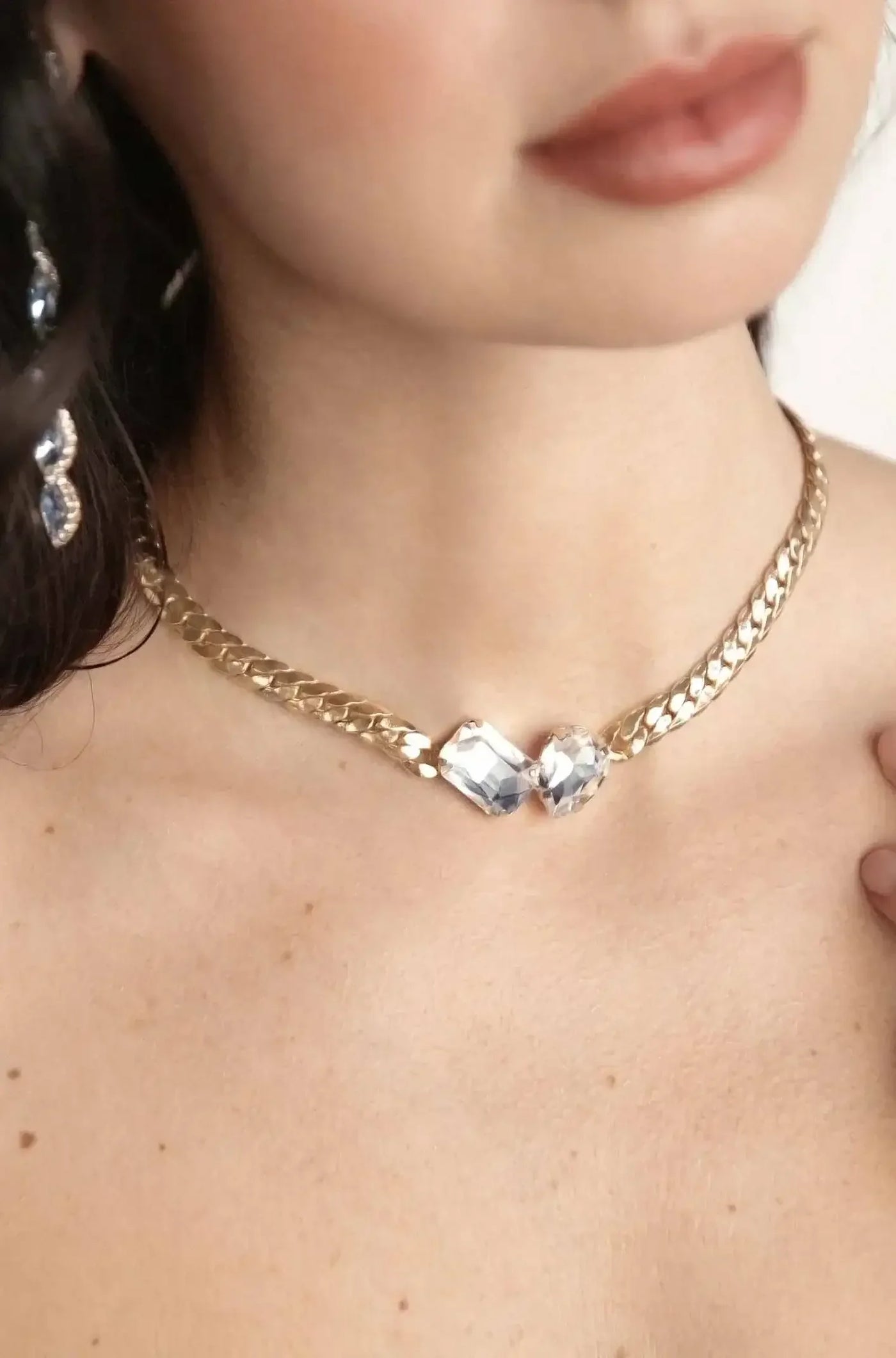 A close-up of a woman's neck adorned with a gold chain necklace featuring two large, clear crystals. She gently touches her collarbone, wearing a black top against a soft background.