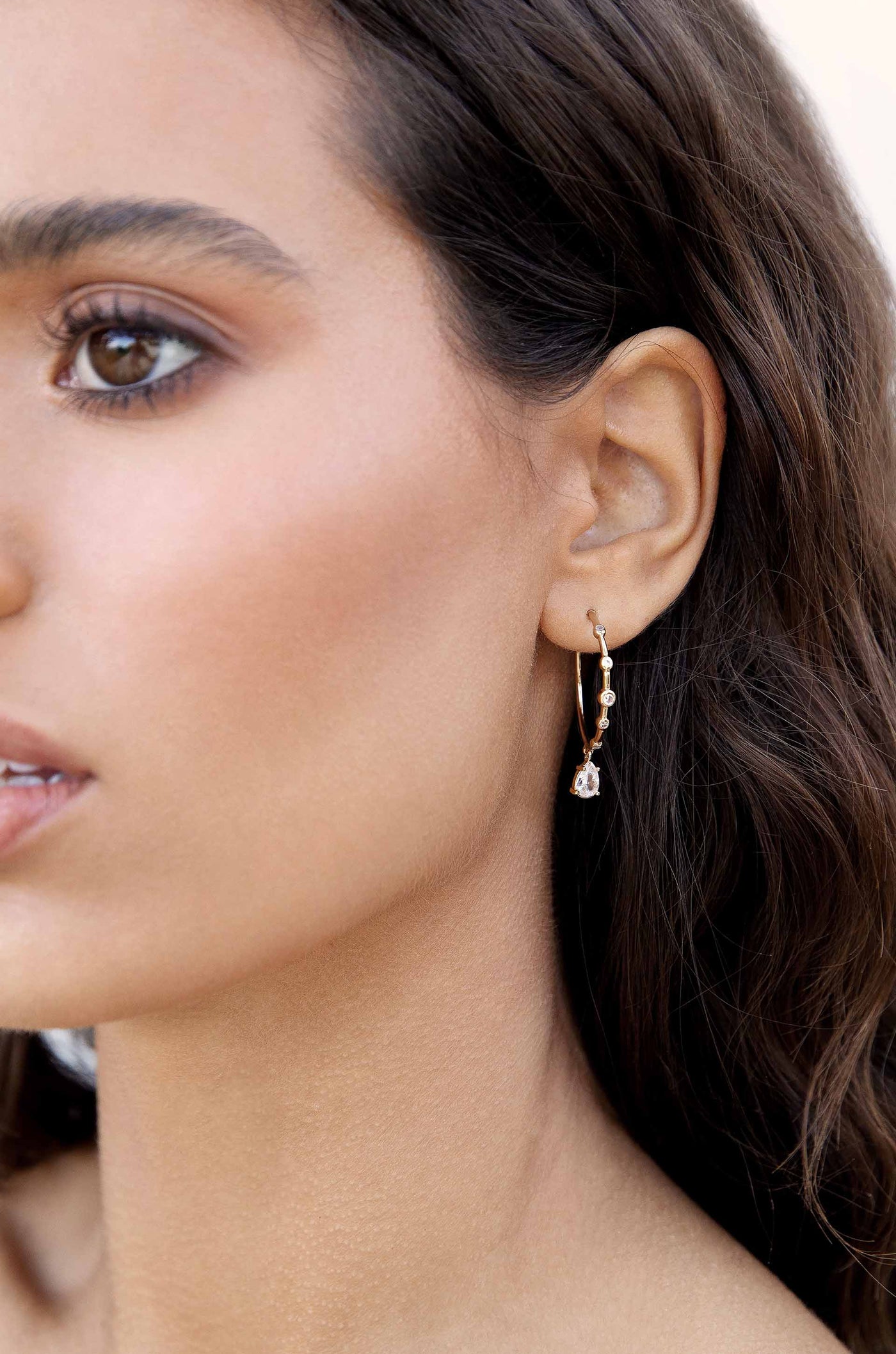 A close-up of a woman's ear adorned with a delicate gold hoop earring featuring small gemstones and a dangling charm, set against a softly blurred neutral background.