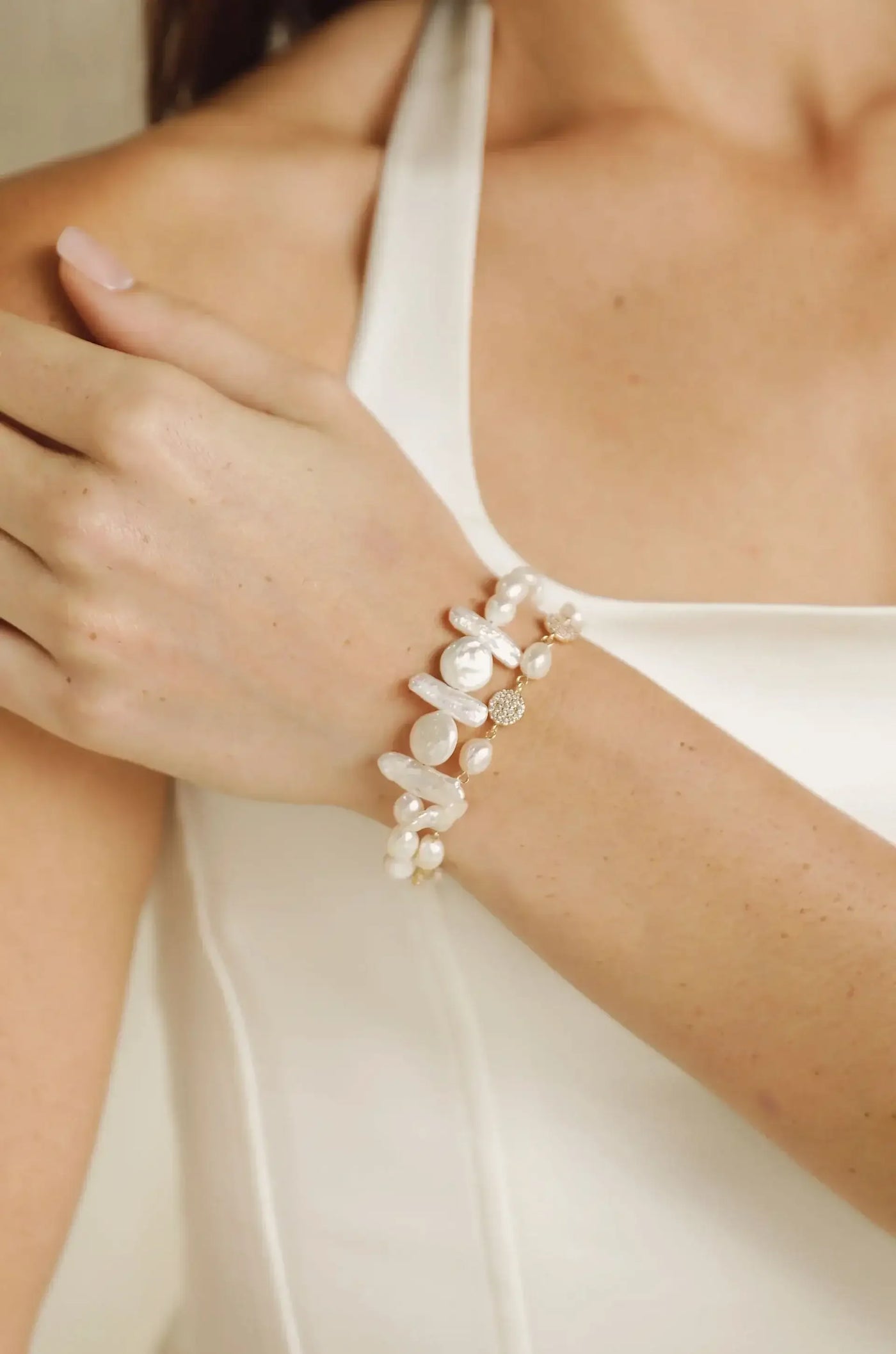A hand adorned with a pearl and gemstone bracelet rests gently on a shoulder. The background features a soft, neutral tone, enhancing the delicate jewelry and skin texture.