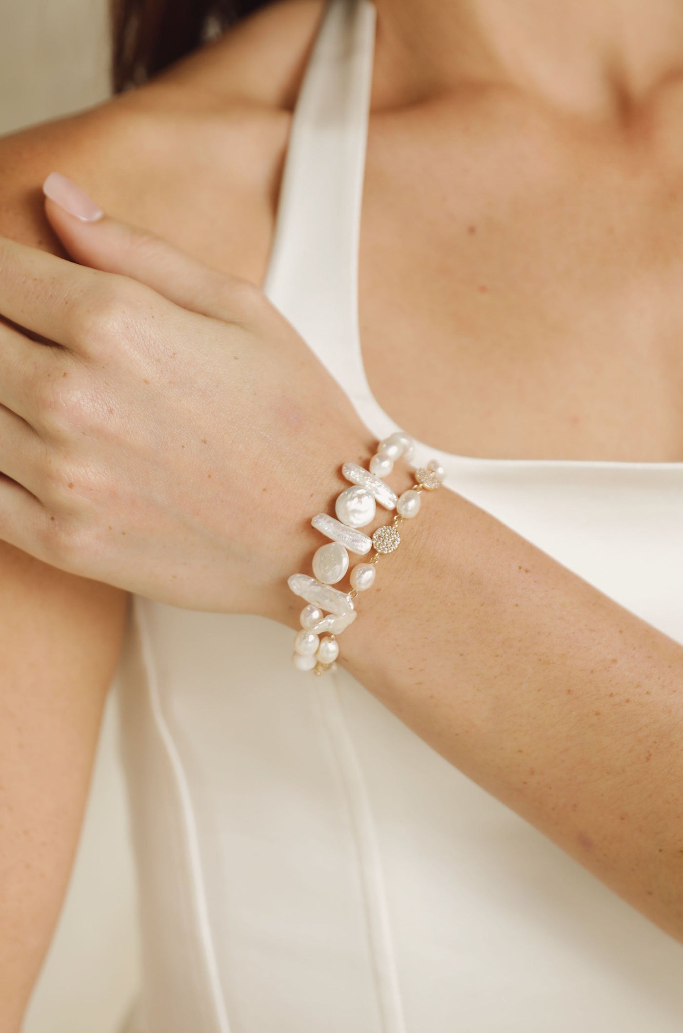 A hand adorned with a pearl and gemstone bracelet rests gently on a shoulder. The background features a soft, neutral tone, enhancing the delicate jewelry and skin texture.