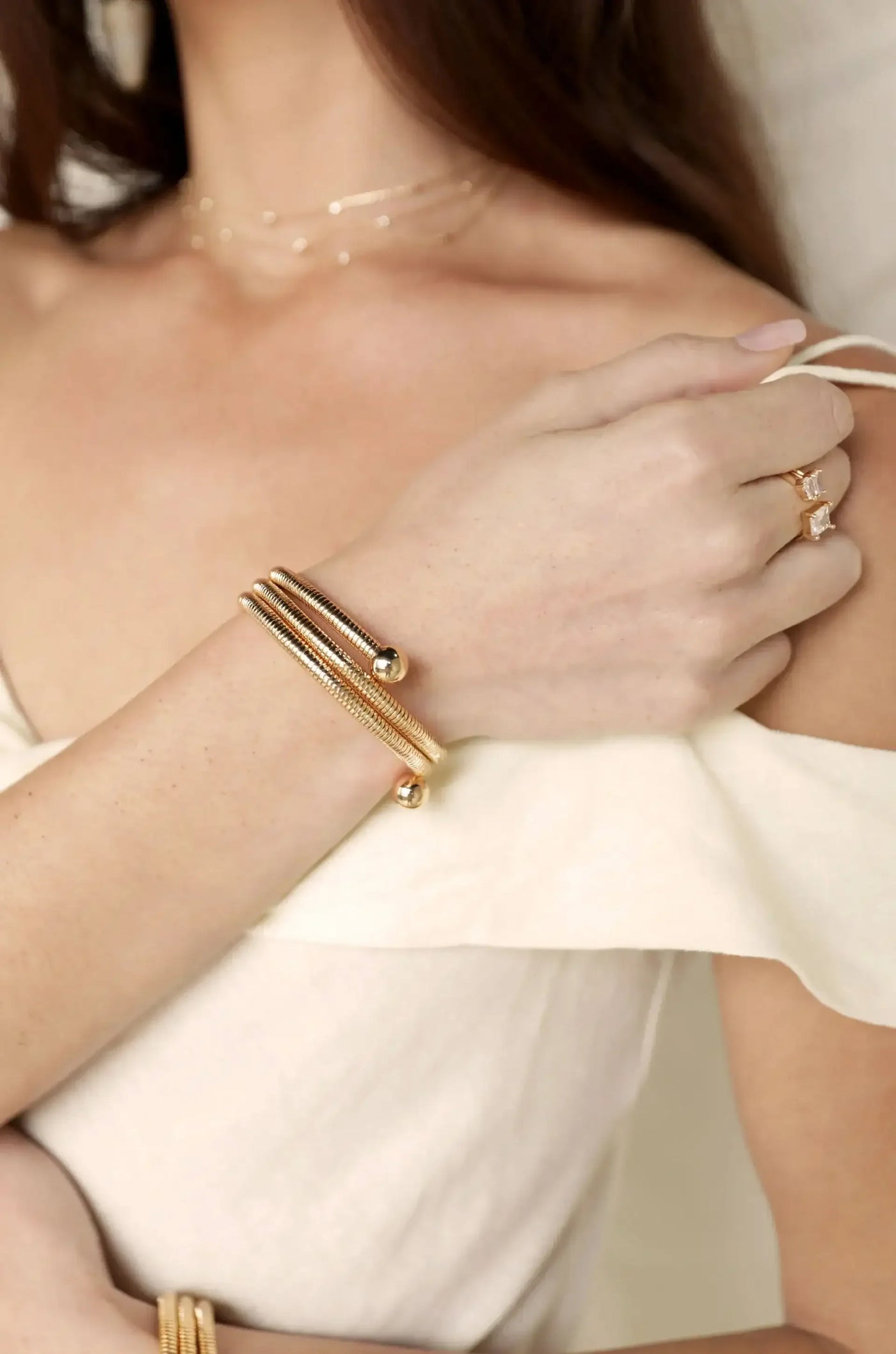 A hand adorned with a multi-stranded golden bracelet rests gently on a bare shoulder, complementing a softly draped, off-white garment in a neutral background.