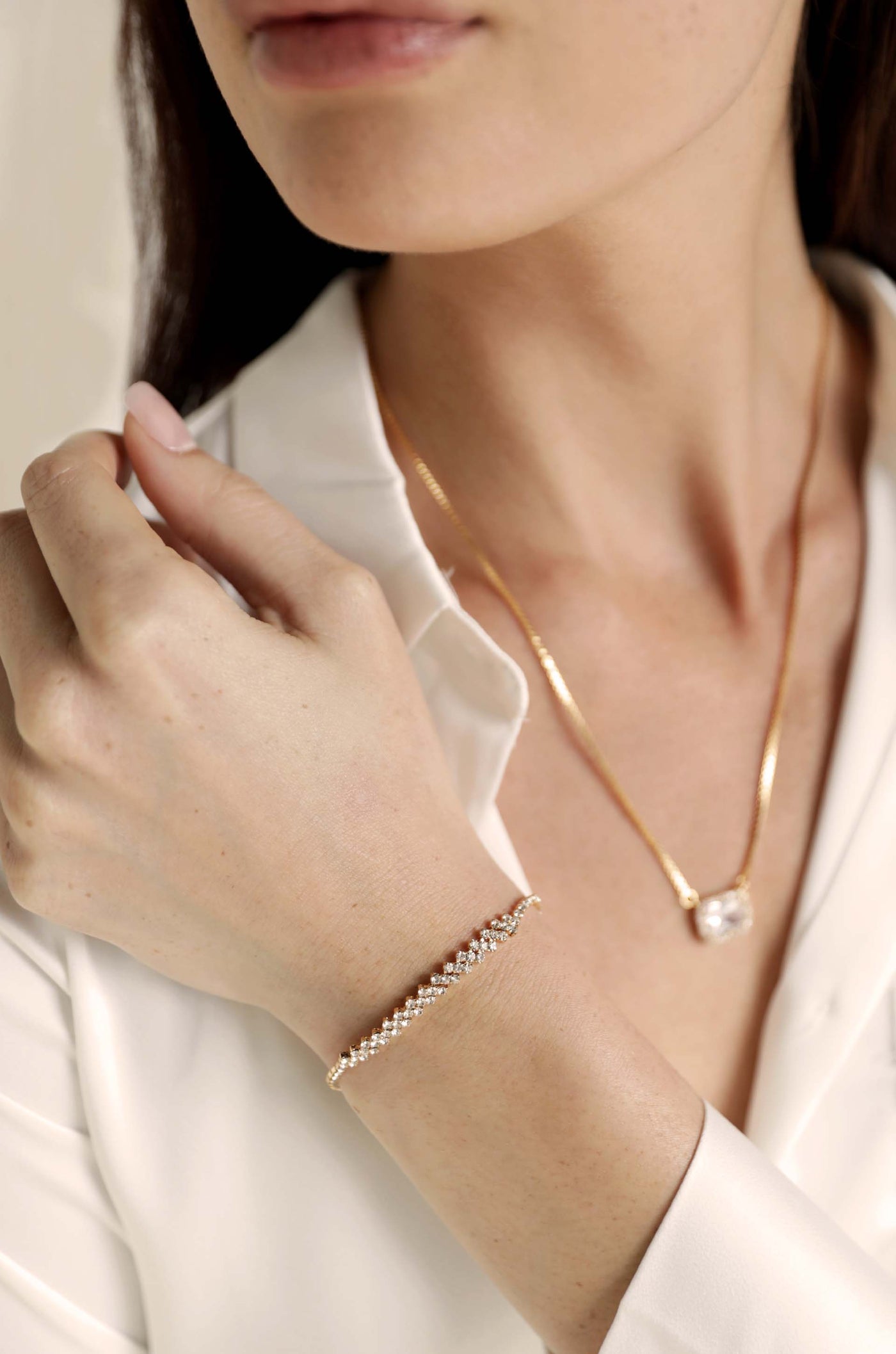 A close-up of a woman's hand gently touching a collar, showcasing a delicate gold bracelet adorned with small gems, set against a soft, neutral background. A gold necklace is also visible.