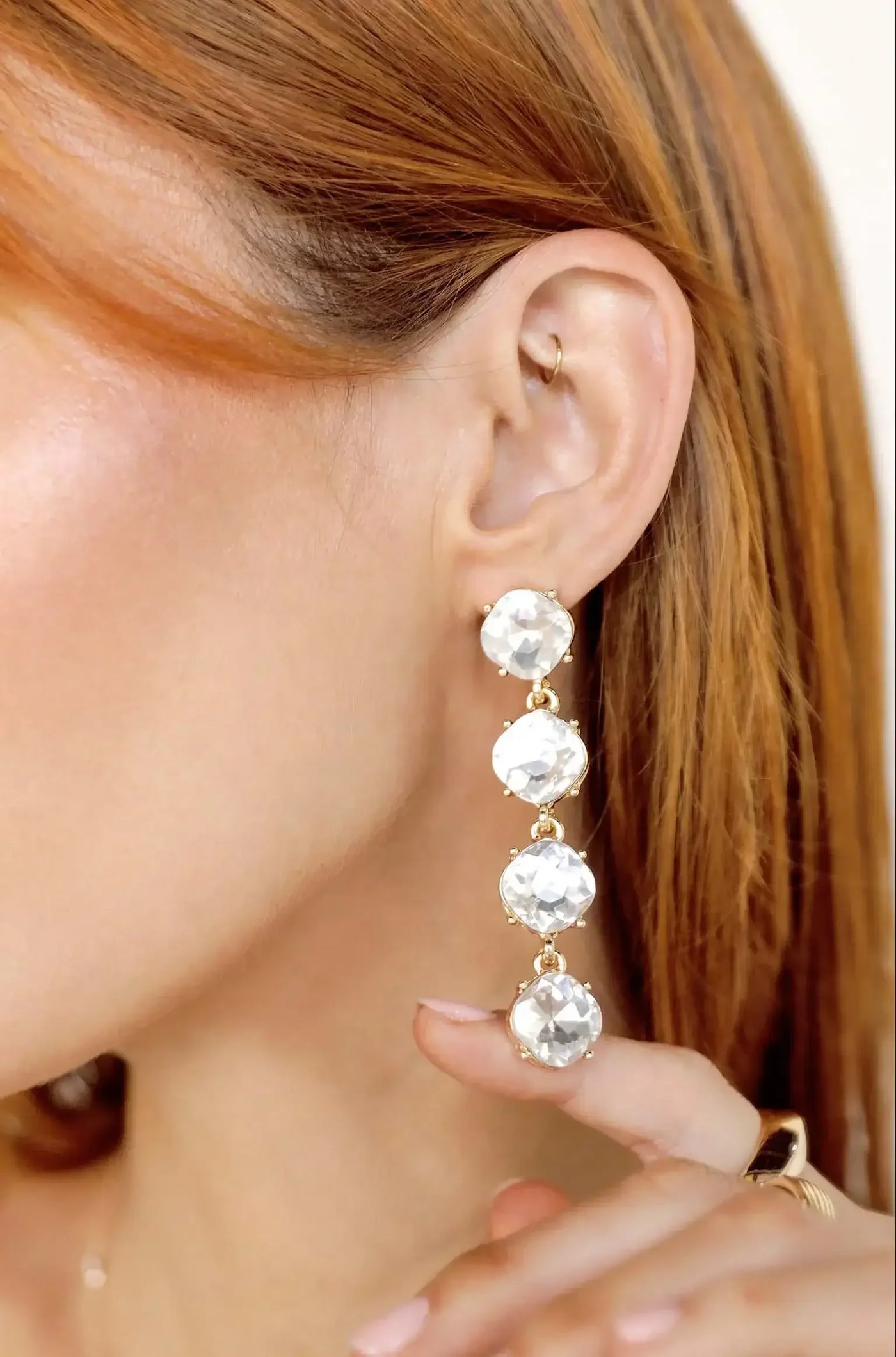 A woman with long, reddish hair wears a sparkling, multi-tiered earring while gently holding it up towards her ear, showcasing the elegant jewelry against a light background.