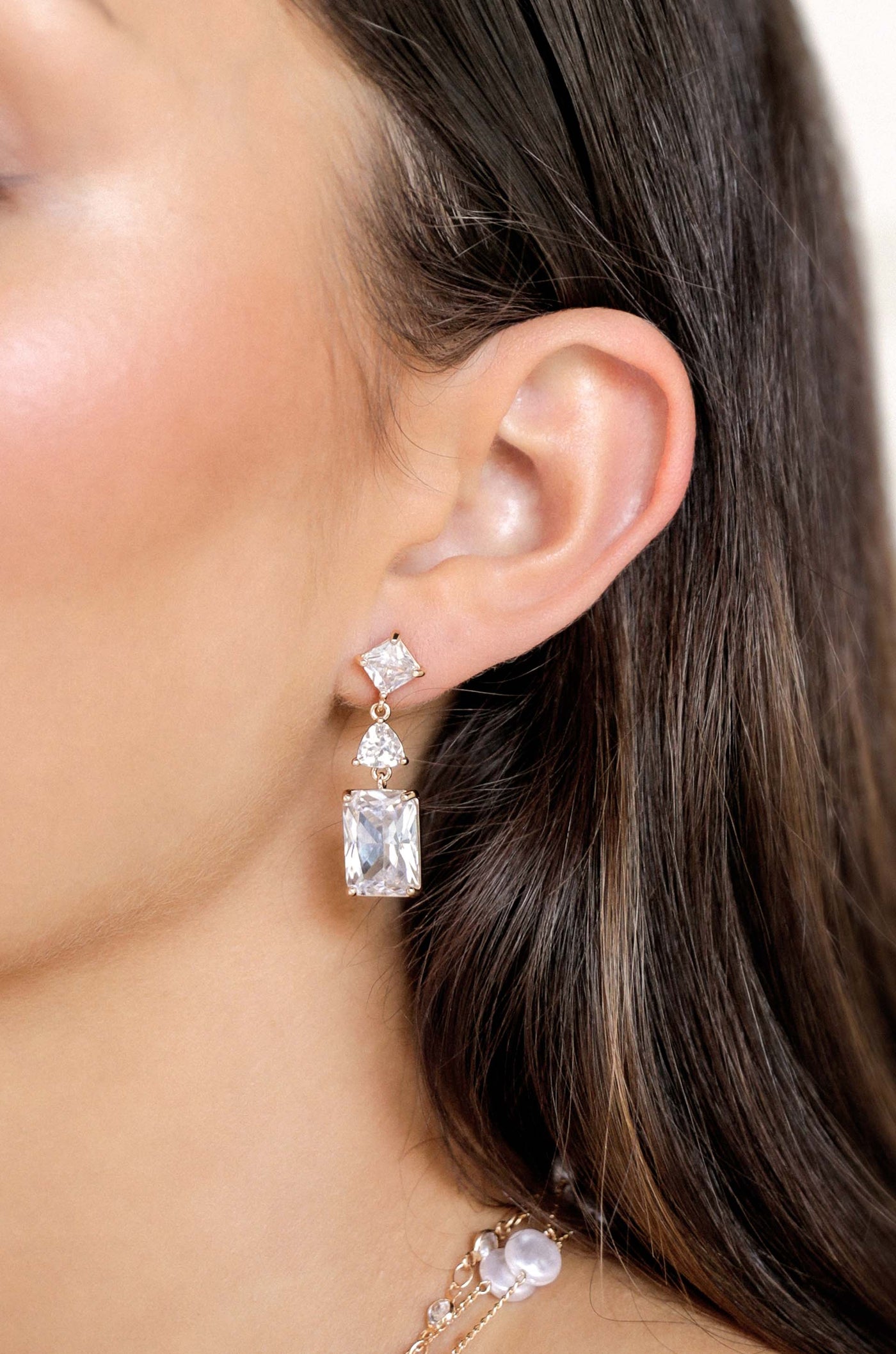 A woman’s ear is adorned with elegant, sparkling drop earrings featuring two large gemstones, one rectangular and one square, set in rose gold, enhancing her side profile against softly flowing hair.