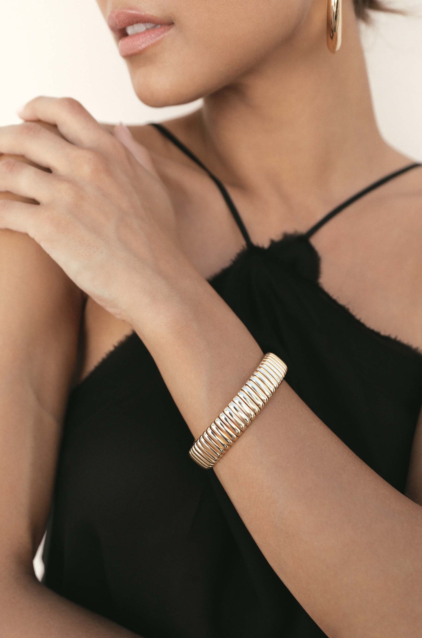 A woman is wearing a gold bangle on her wrist while gently resting her hand on her shoulder, dressed in a black sleeveless top against a light, neutral background.