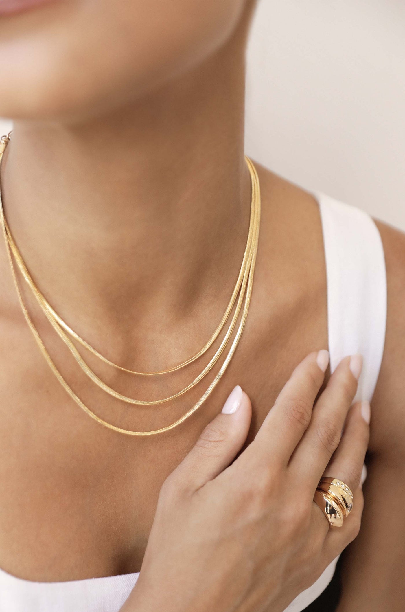 A layered gold necklace rests elegantly on a neck, while a hand gently touches the jewelry. The background is softly blurred, emphasizing the accessories and skin tone against a neutral setting.