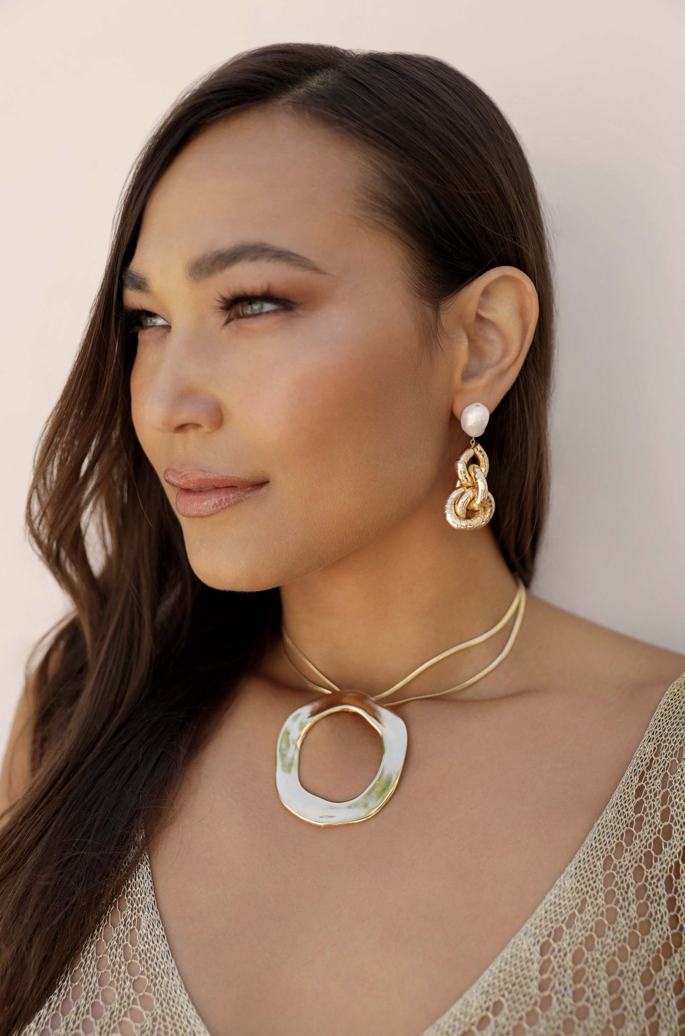 A woman with long, dark hair wears distinctive jewelry: a large, circular pendant necklace and ornate earrings. She gazes thoughtfully to her side against a neutral background.