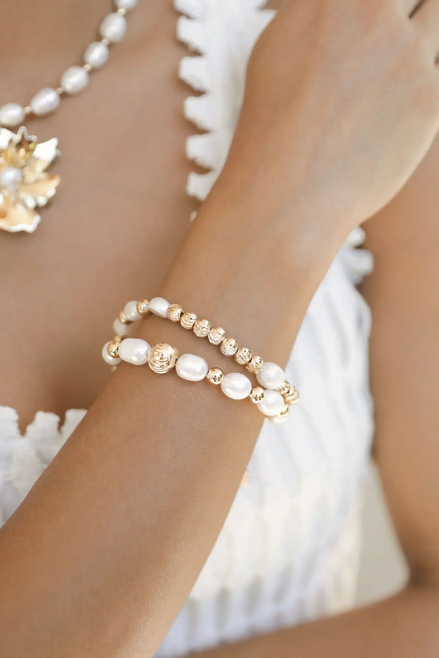 A woman’s arm adorned with two elegant bracelets, featuring a combination of pearls and gold accents. The background is softly blurred, highlighting the jewelry against a light-colored clothing texture.