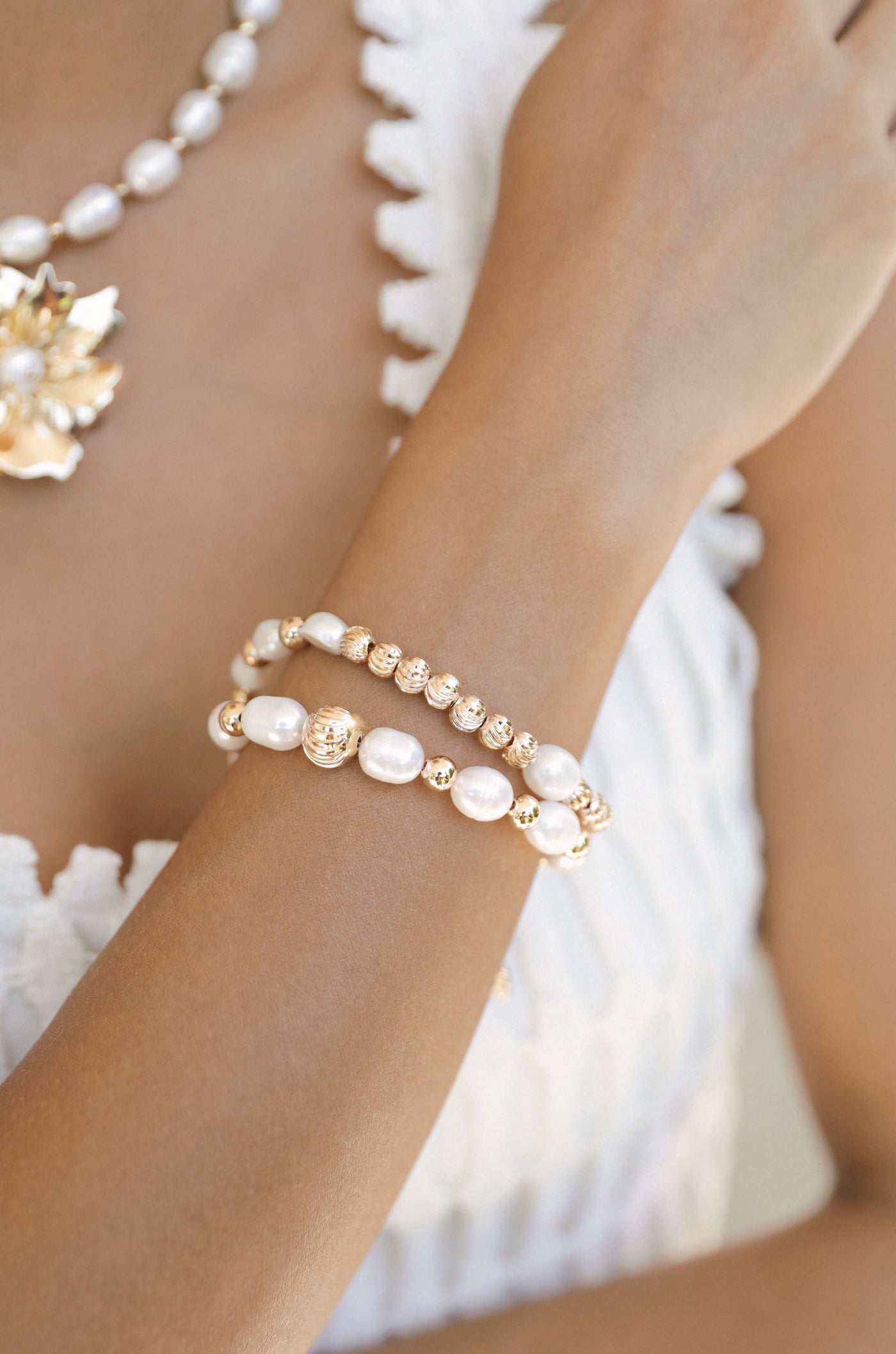 A woman’s arm adorned with two elegant bracelets, featuring a combination of pearls and gold accents. The background is softly blurred, highlighting the jewelry against a light-colored clothing texture.