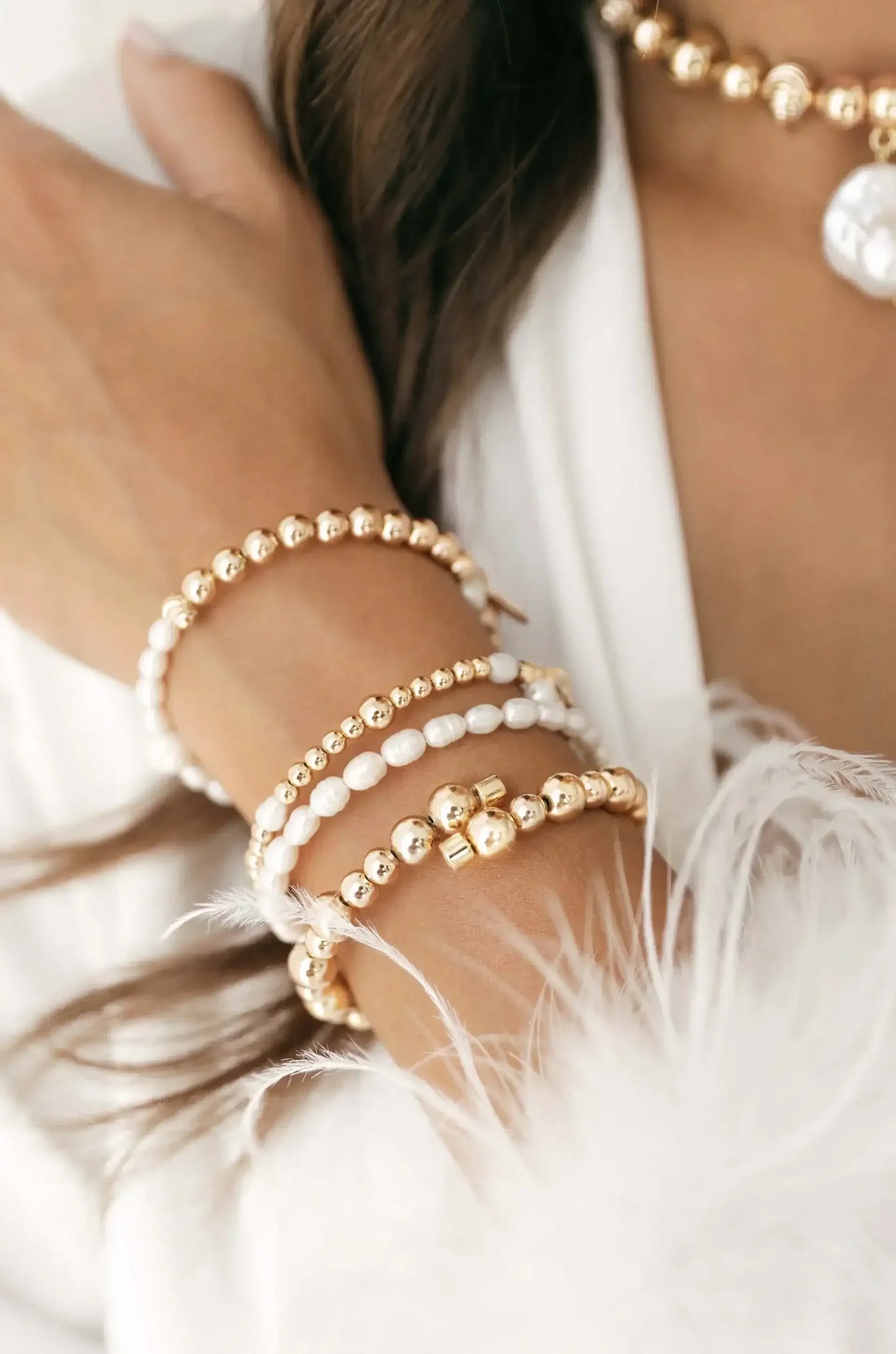 A hand adorned with multiple elegant bracelets made of gold and white beads rests against a soft, white feathered fabric, suggesting a stylish and luxurious setting.