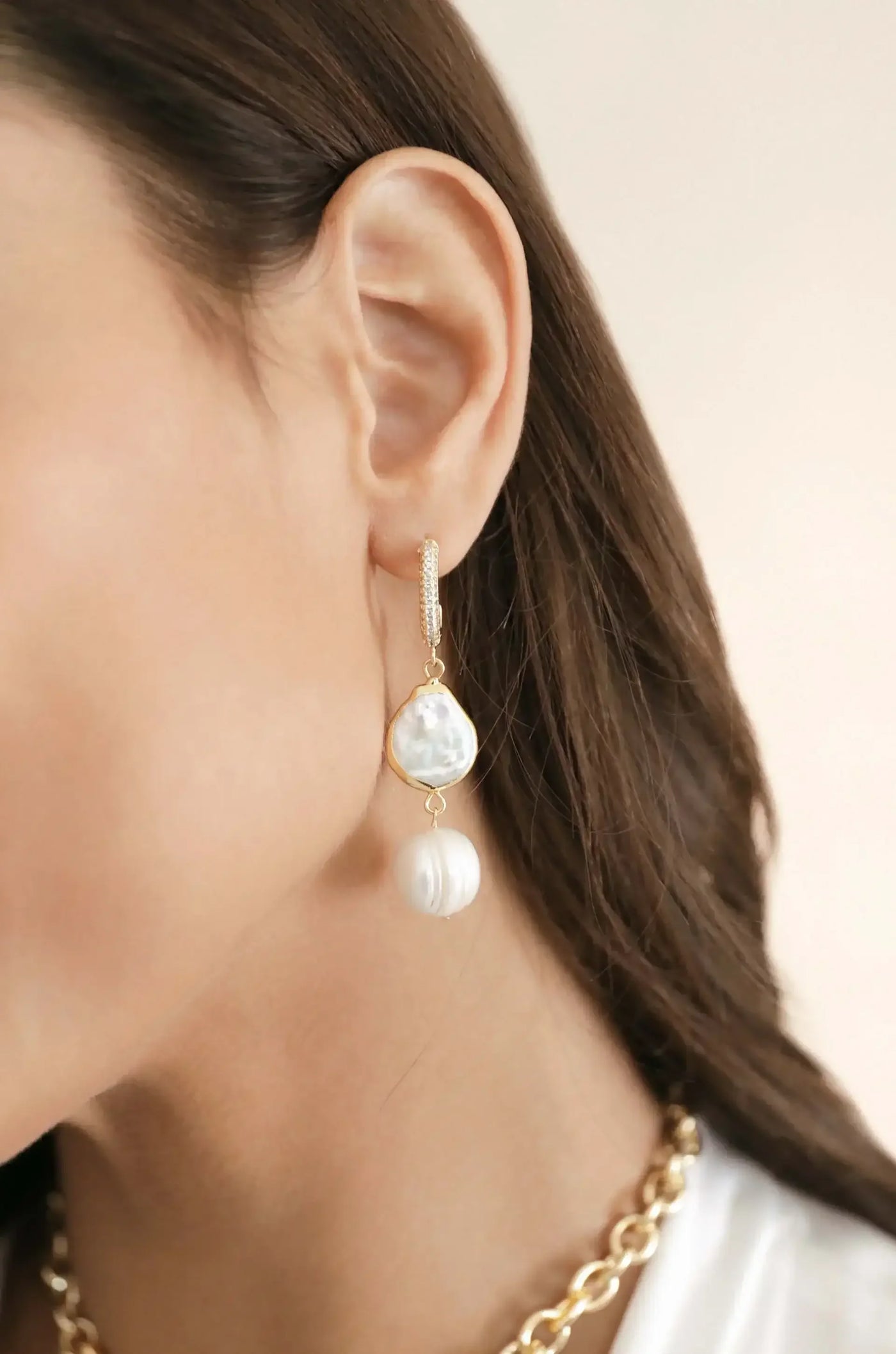 A close-up of a woman's ear adorned with elegant dangling earrings featuring a large, irregularly shaped pearl and a smaller round pearl, set against a soft, neutral background.