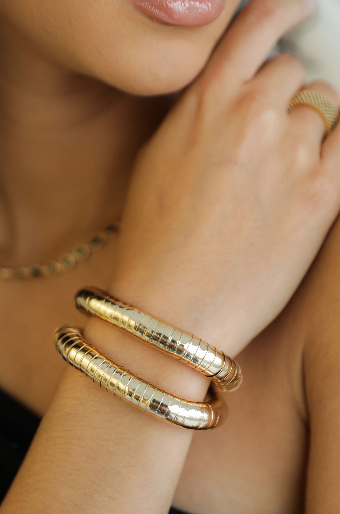 A pair of shiny metallic bangles encircles a woman's wrist while her fingers lightly touch her arm. She has glossy lips and wears minimal jewelry against a softly blurred background.