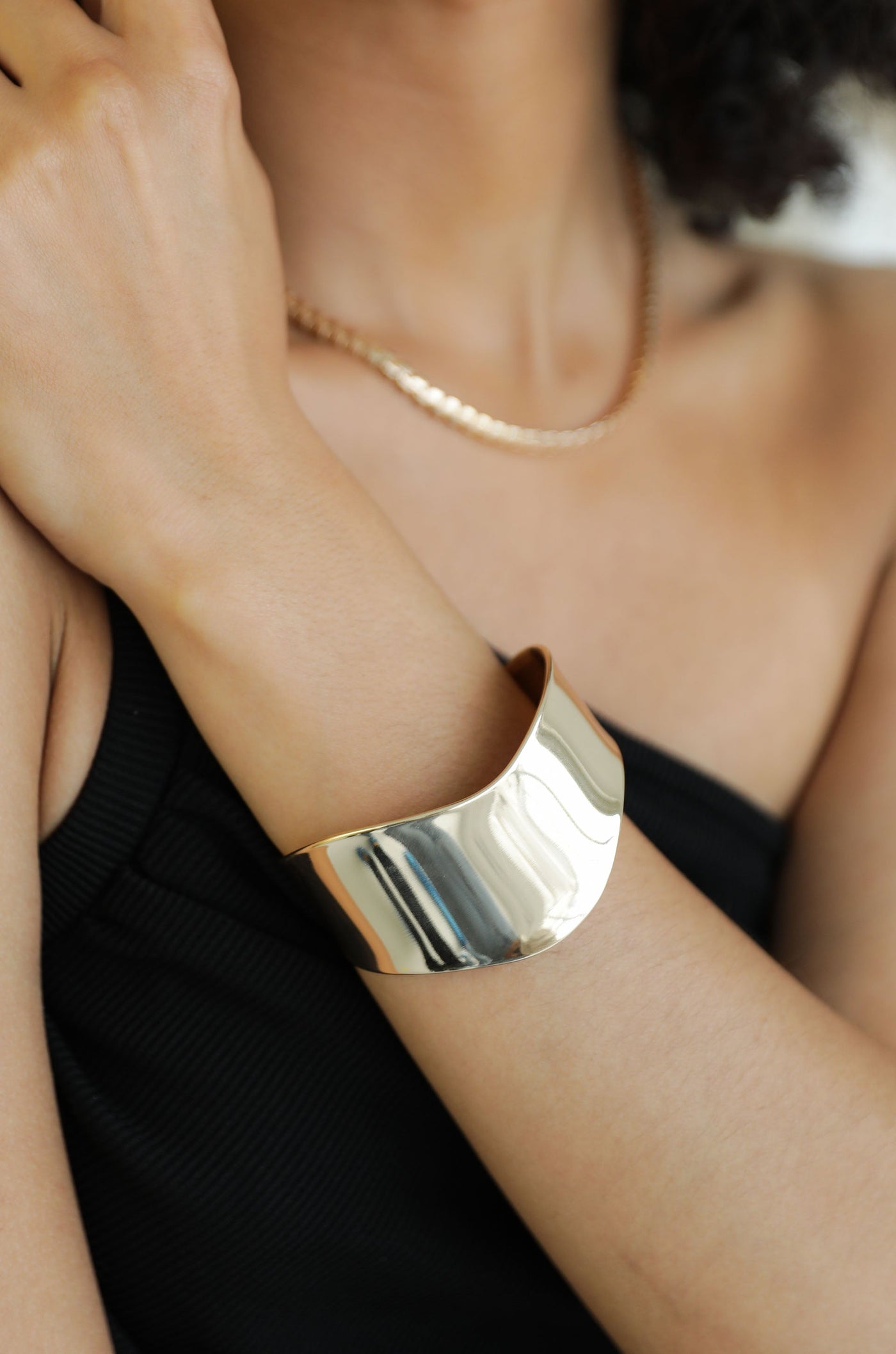 A gold cuff bracelet is elegantly positioned on the wrist of a person, enhancing a minimalistic black outfit. The arm is slightly bent, showcasing the bracelet's shiny, curved design against soft skin.
