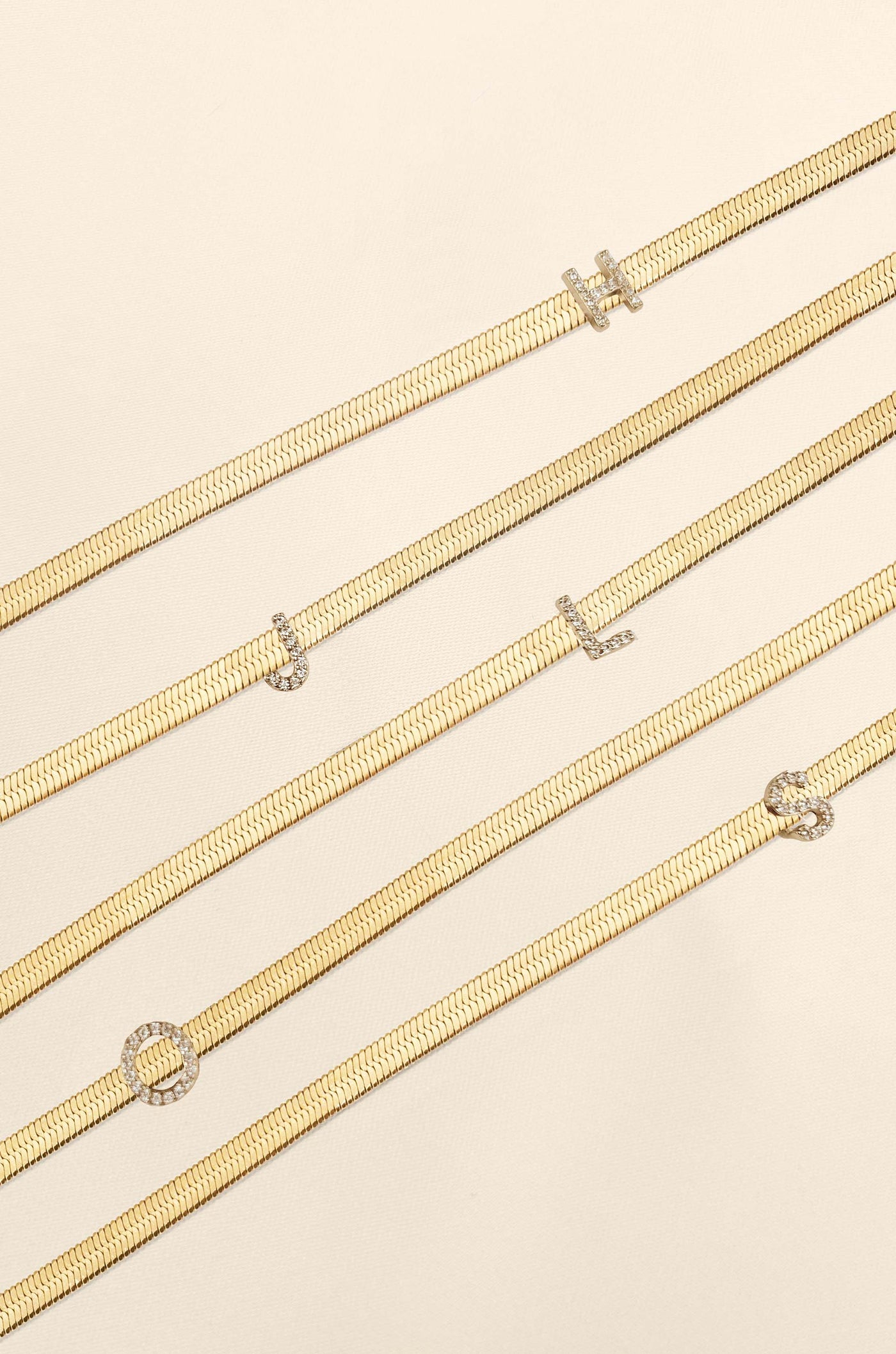 Five gold necklaces with various decorative elements lay flat on a soft, light beige surface. They feature letter-shaped embellishments, creating a personalized design.