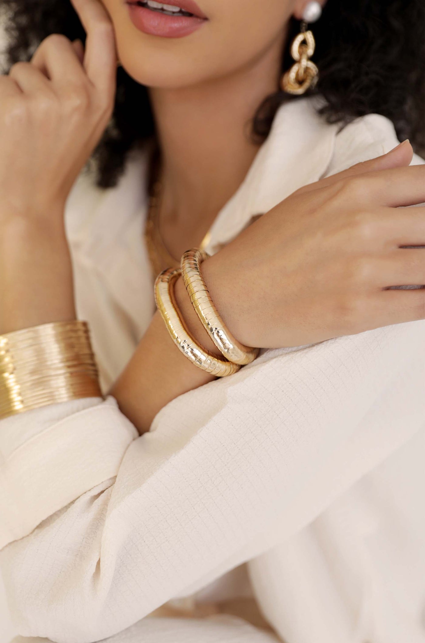 Three gold bangles adorn a woman's wrist as she ponders thoughtfully, resting her finger on her chin, while wearing a light-colored, textured shirt in a softly lit environment.