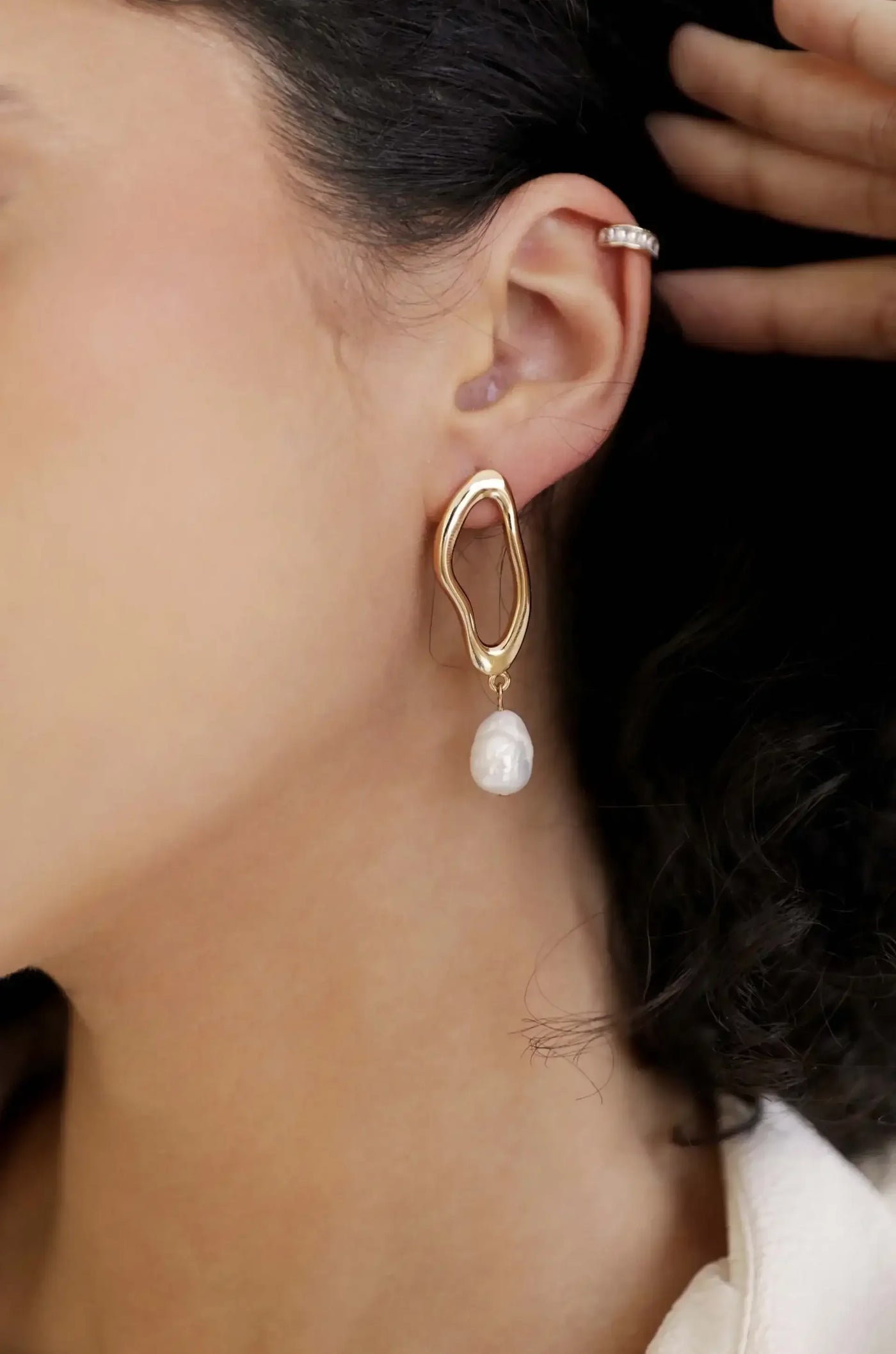 A gold earring with an abstract shape hangs from the ear of a woman, featuring a dangling pearl. The setting is softly lit, enhancing her natural features and highlighting the earring's design.