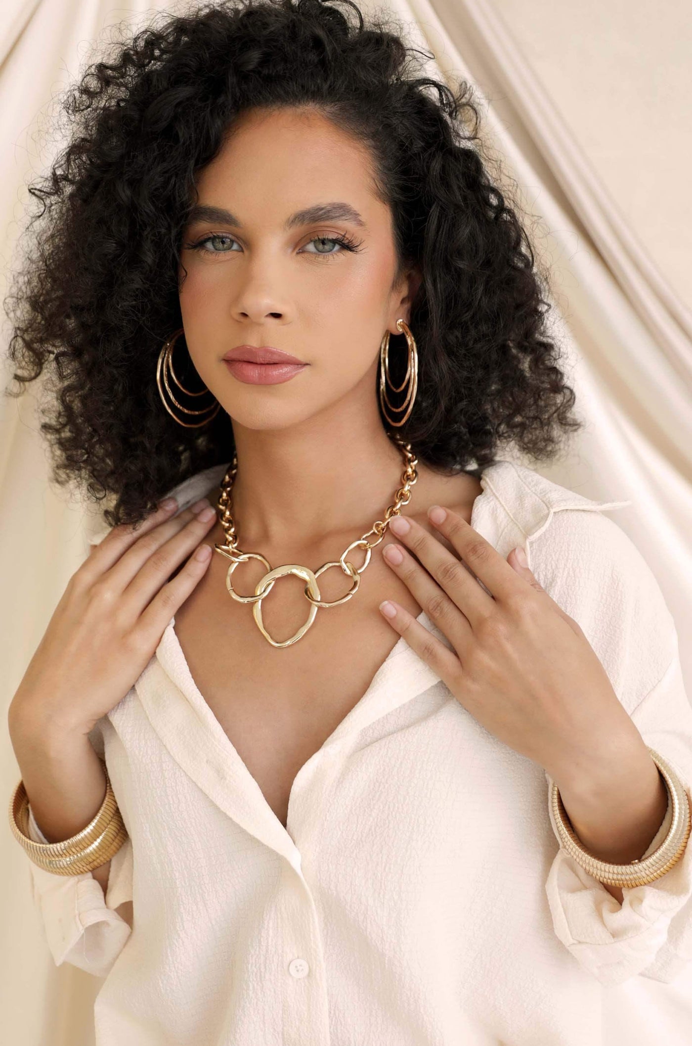 A woman with curly hair wears bold hoop earrings and a statement necklace while gently touching her collar. She is dressed in a white button-down shirt, against a soft, neutral backdrop.