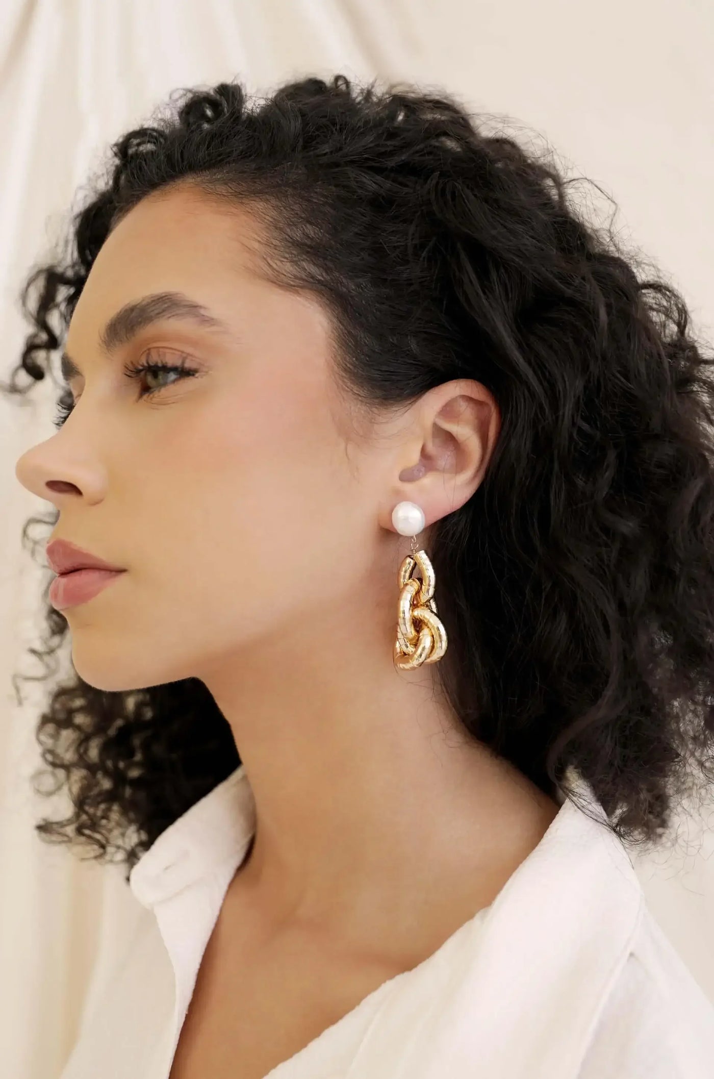 A woman with curly hair wears a large, gold chain-style earring and a pearl stud on her left ear, set against a soft, neutral background.