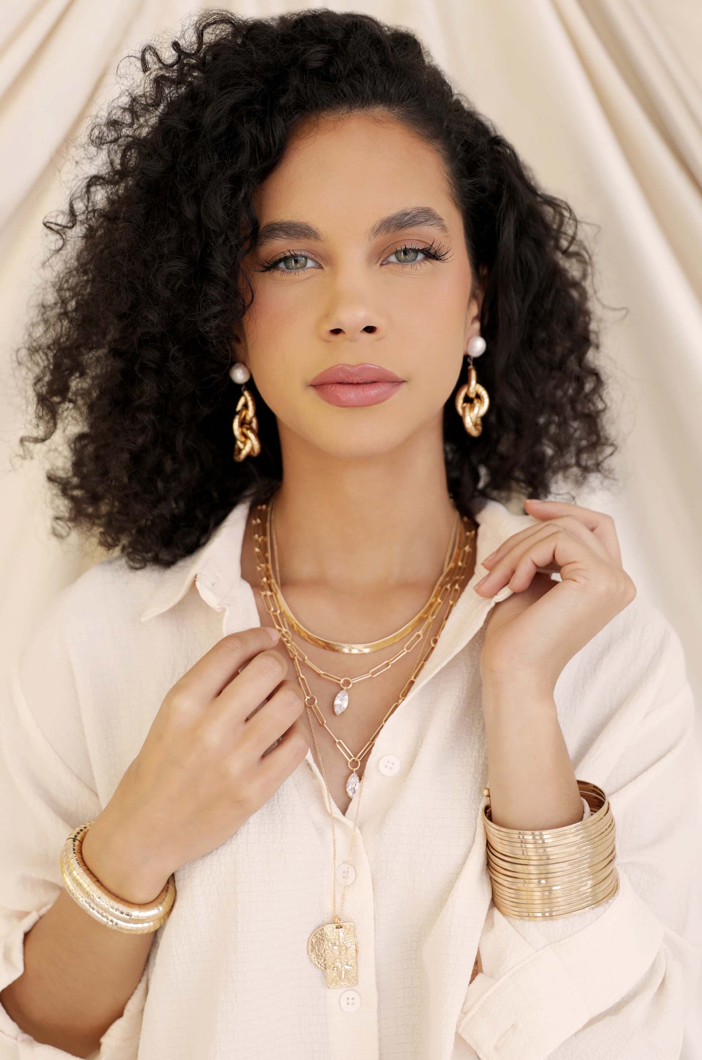 A woman with curly hair wears multiple gold necklaces and bracelets, posing gracefully. The light-colored, soft fabric backdrop enhances her elegant look against a neutral setting.