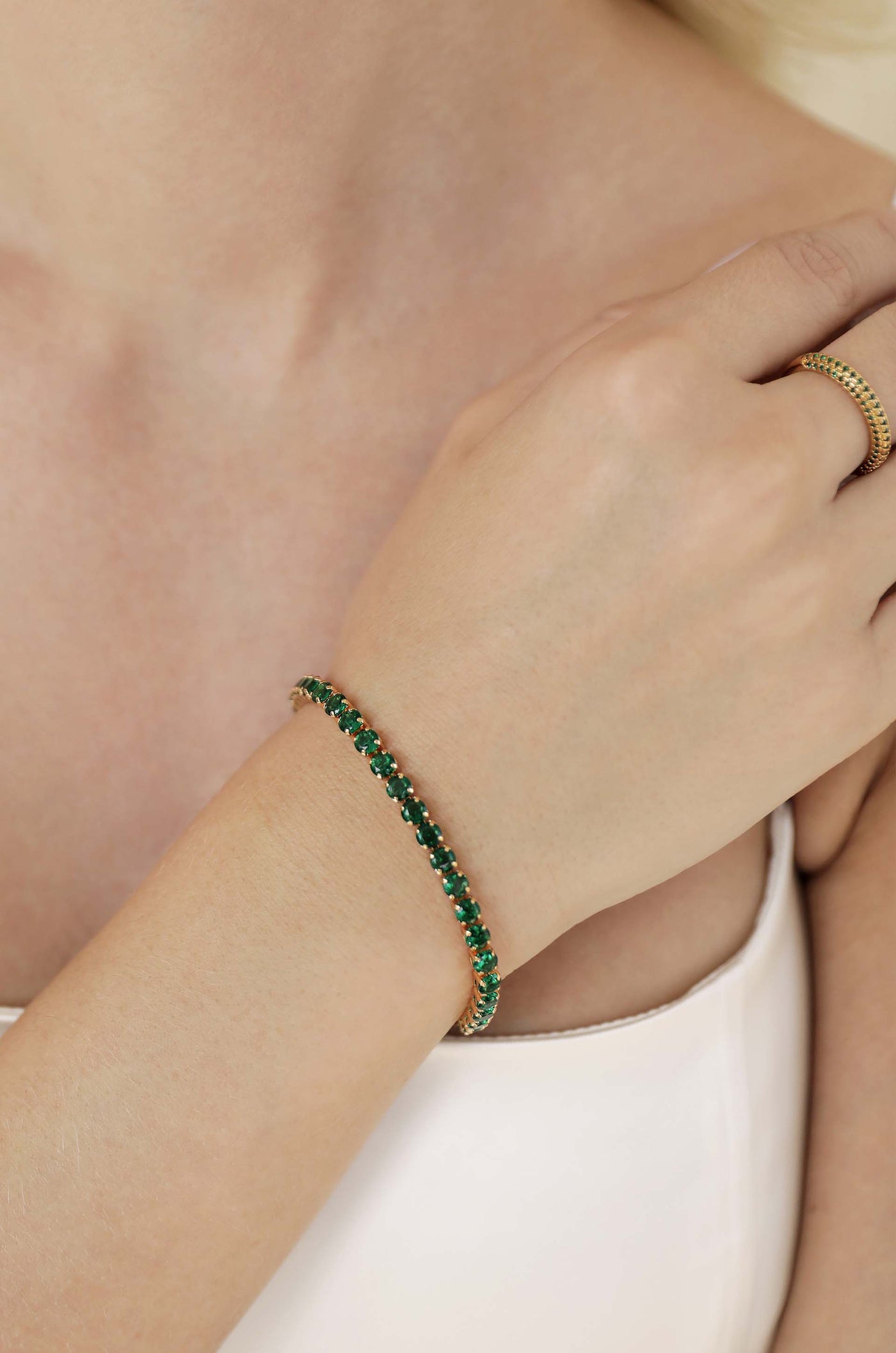 A delicate gold bracelet set with green gemstones adorns a woman's wrist. She gently touches her arm, standing against a soft, neutral background that enhances the jewelry's elegance.