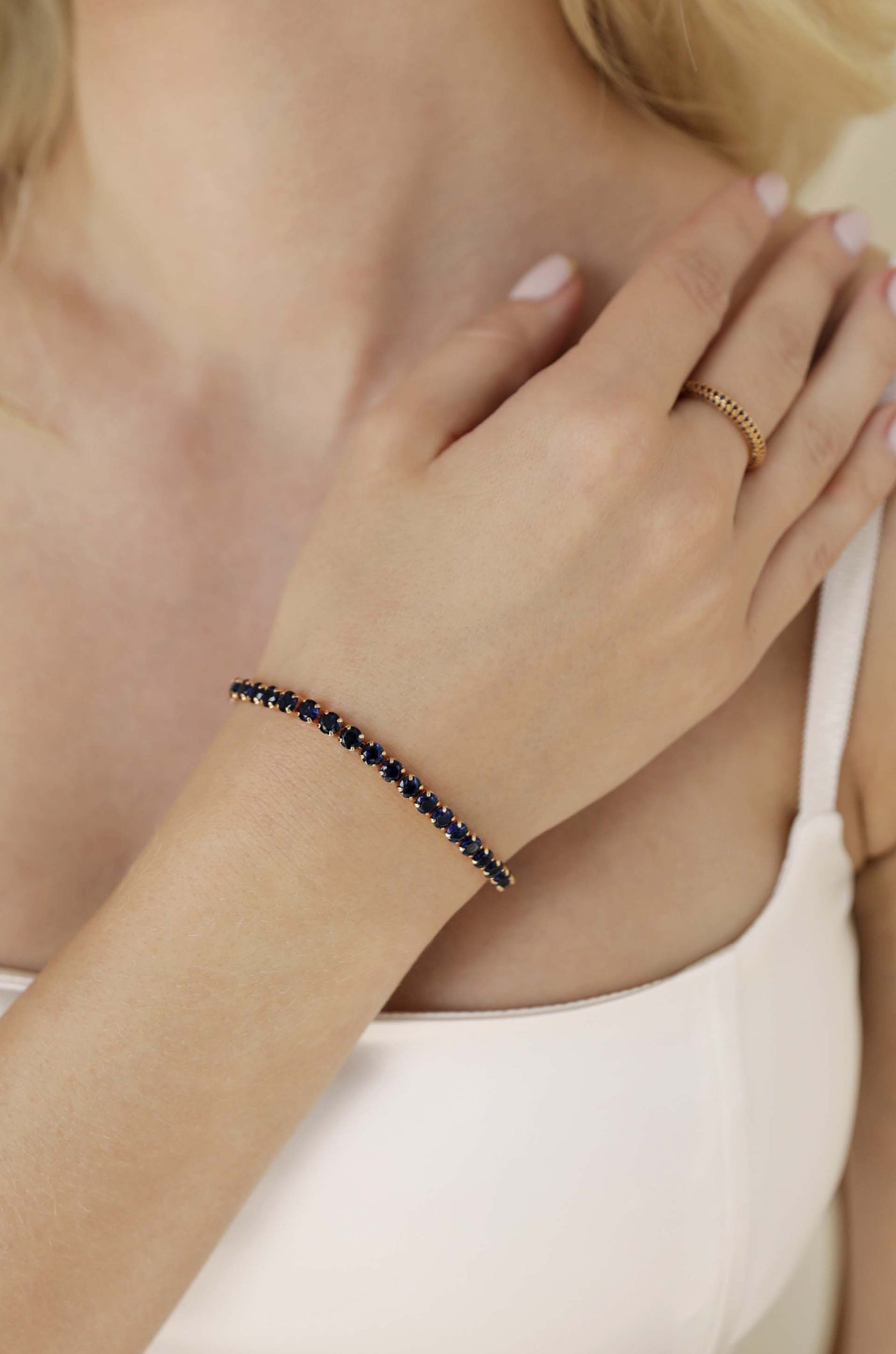 A hand adorned with a delicate gold bracelet featuring dark blue stones rests on a bare shoulder. The background is soft and neutral, highlighting the jewelry and skin tone.