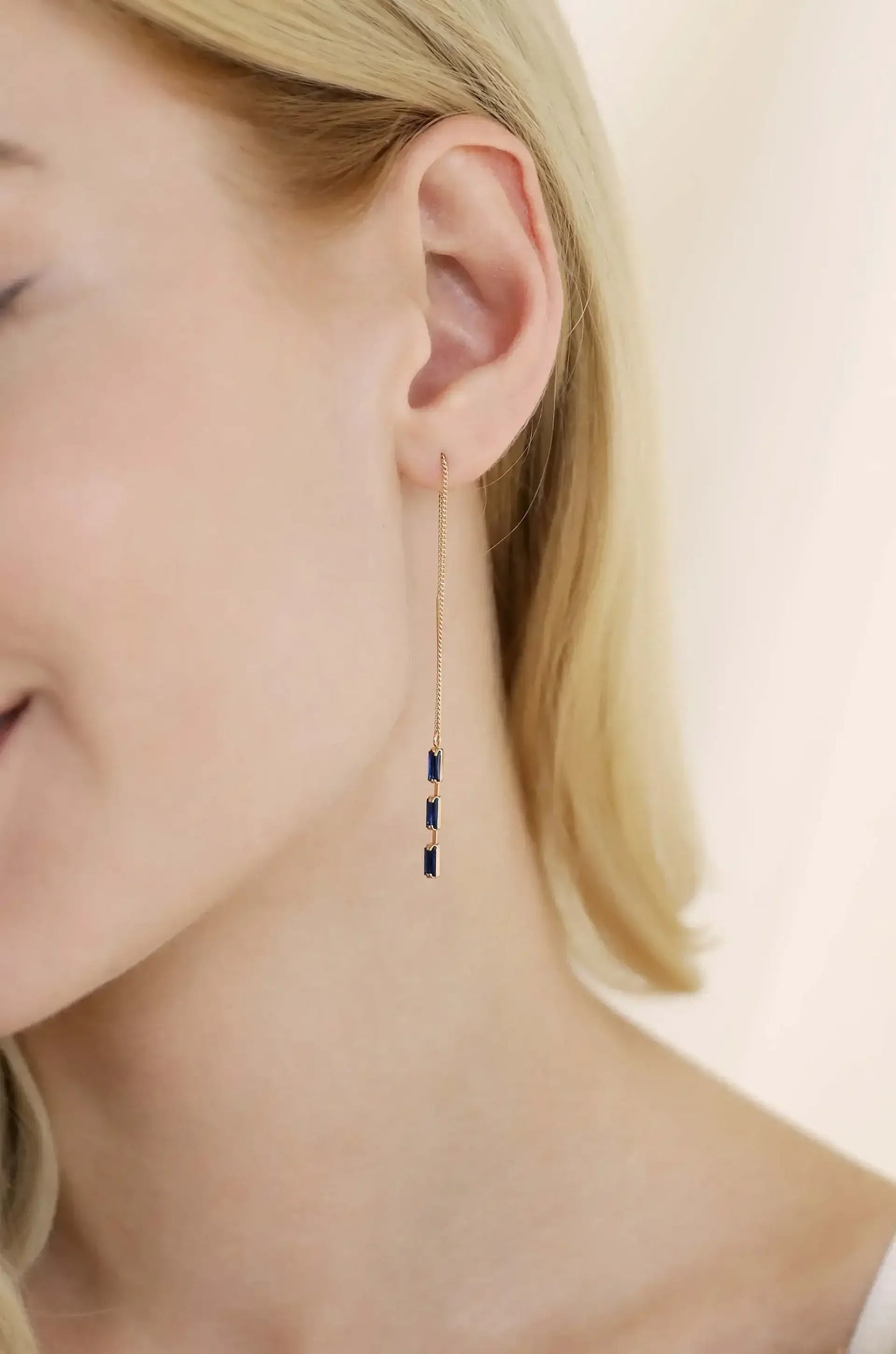 A dangling earring adorned with multiple small, dark blue gemstones hangs from a woman’s ear. She has blonde hair and is smiling softly against a softly lit, neutral background.
