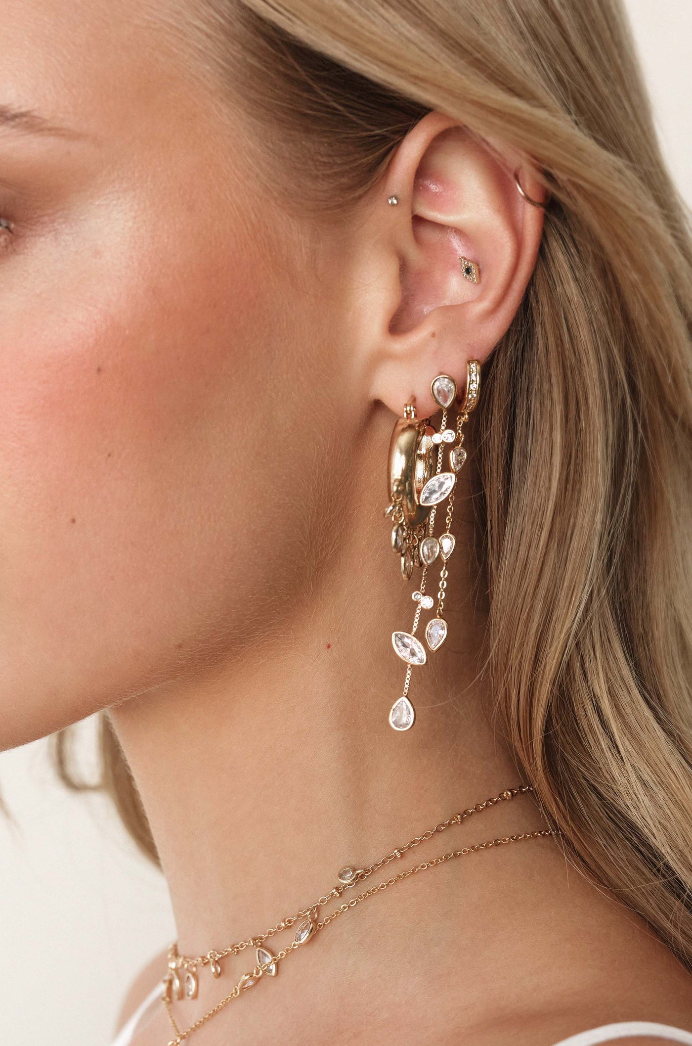 A woman’s ear showcases a collection of elaborate gold earrings featuring dangling gemstones and chains, illuminated by soft light, highlighting the intricate design against her smooth skin.