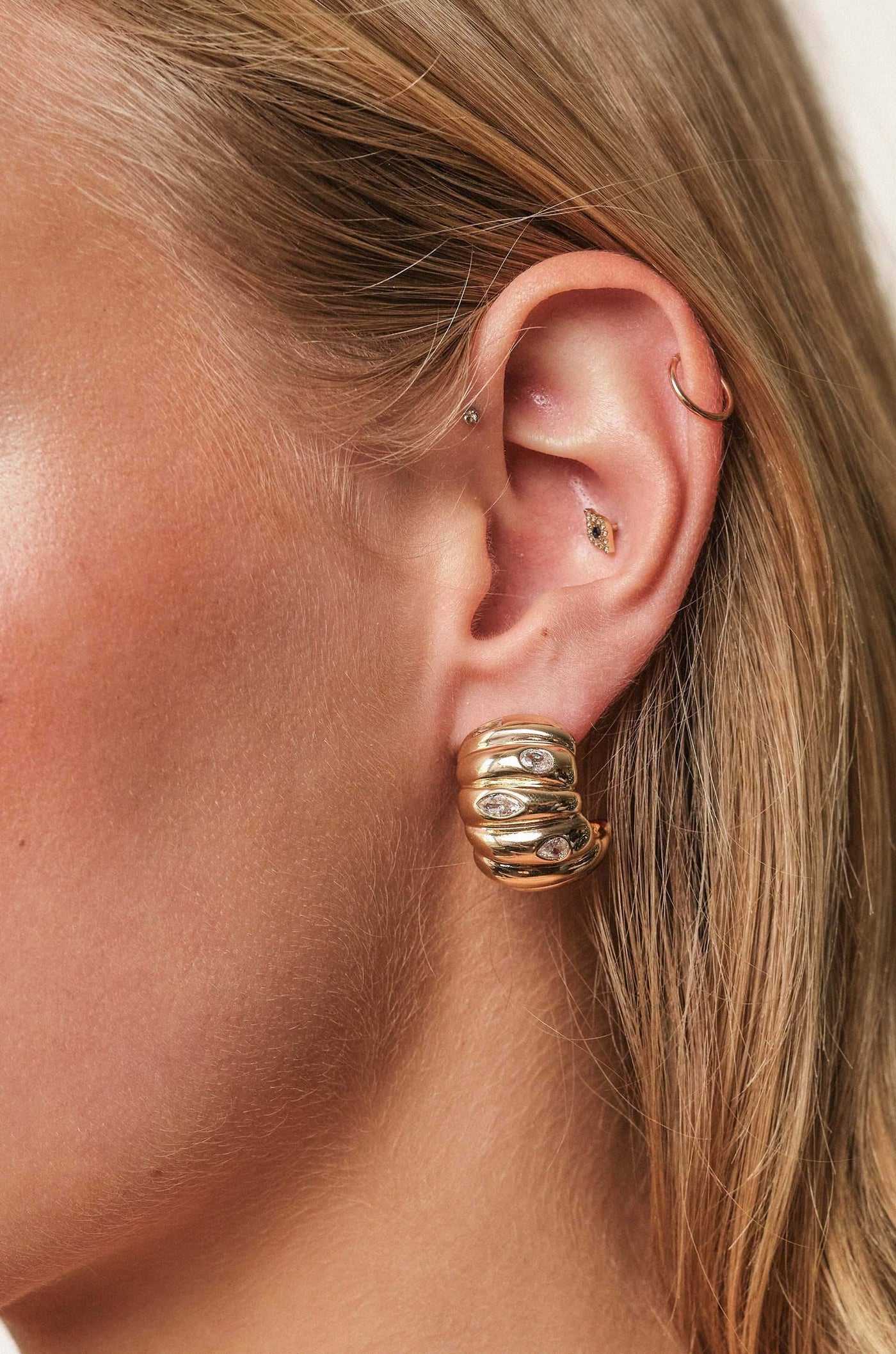 A close-up of a woman's ear showcases a large, spiral gold earring adorned with small diamonds, highlighting delicate piercings and soft blonde hair in a neutral background.