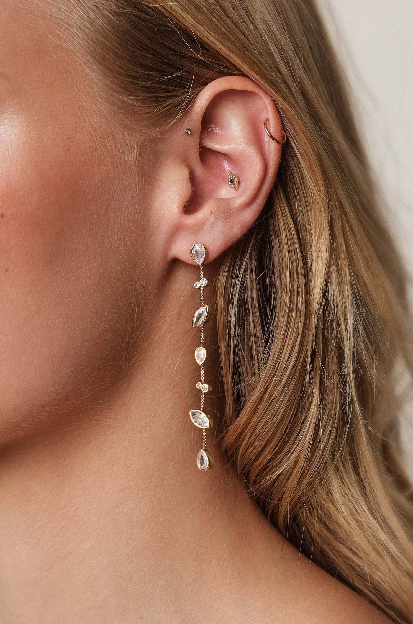 A silver earring with dangling gemstones is adorning the lobe of a person's ear. The hair is styled loosely, framing the face against a neutral background.