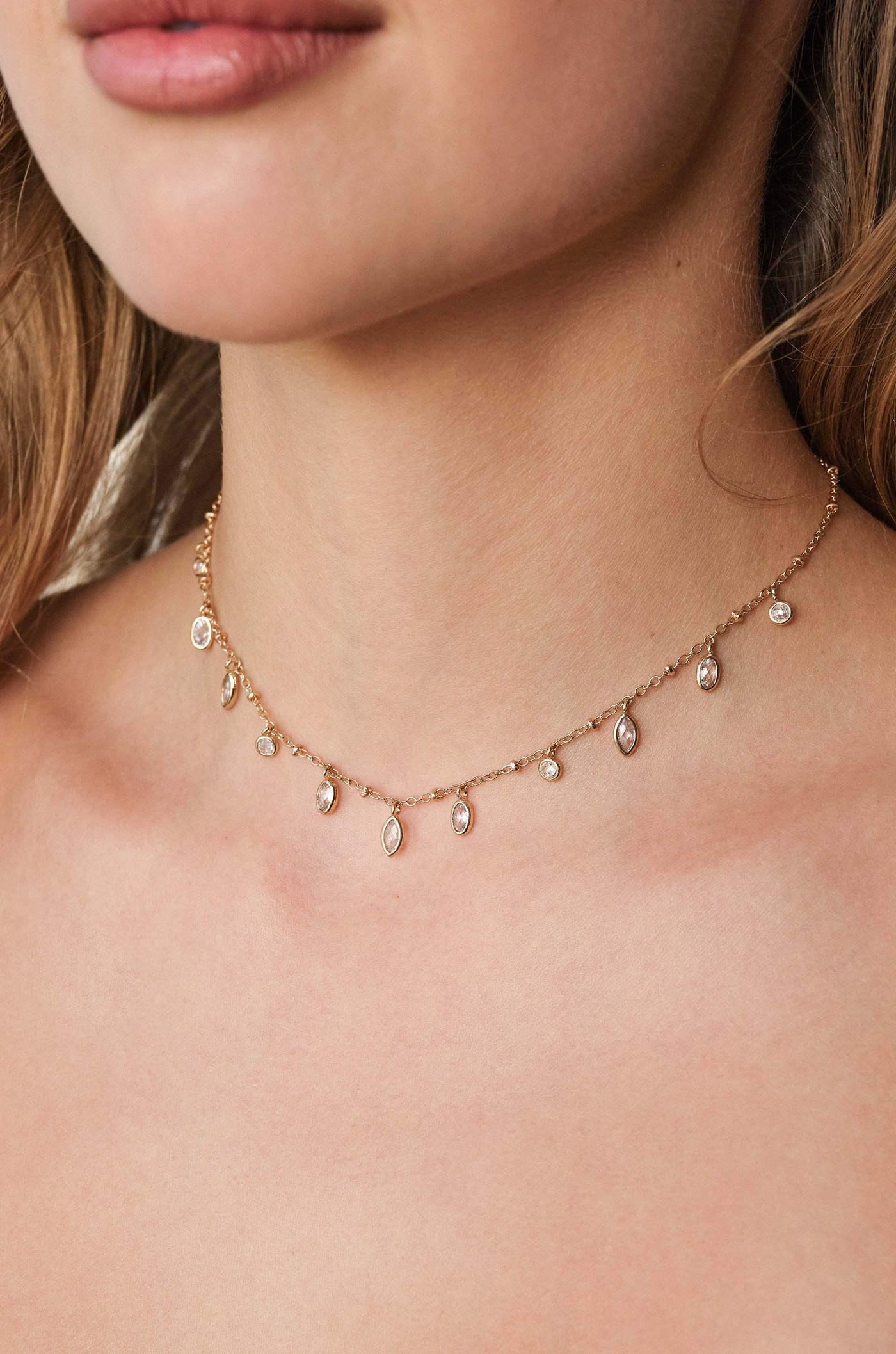 A delicate gold necklace adorned with small, sparkling gems rests on a woman's neck, accentuating her skin. The design features a series of dangling charms that catch the light.