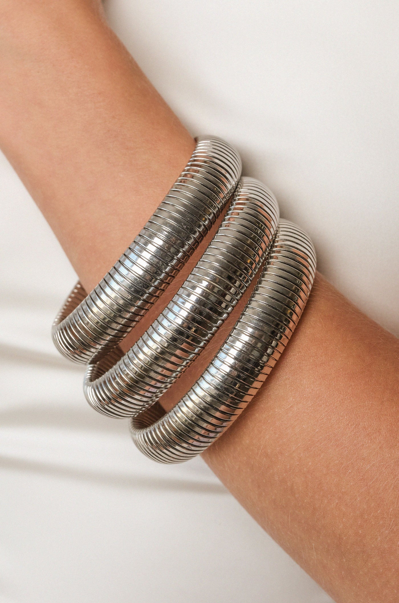 A metallic bangle, consisting of three stacked, ribbed bands, adorns a light-skinned arm. The background is neutral, emphasizing the shiny texture of the jewelry.