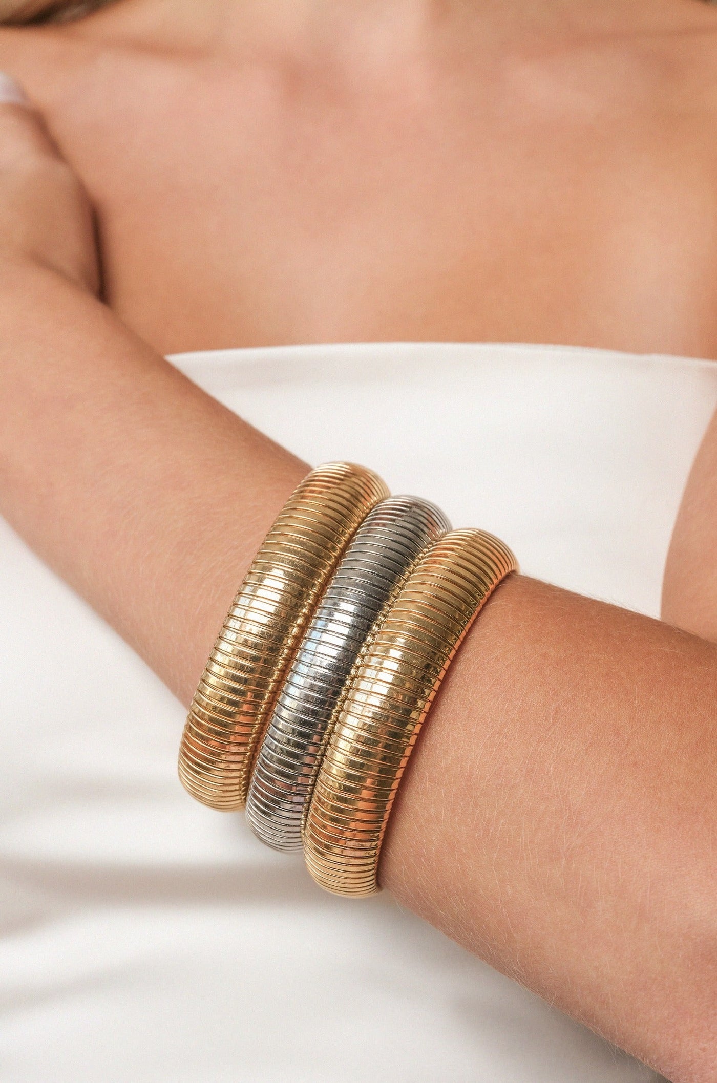 Three metallic bangles—two gold and one silver—are stacked on a bare arm. The background features a soft, neutral tone, emphasizing the jewelry's shine and texture.