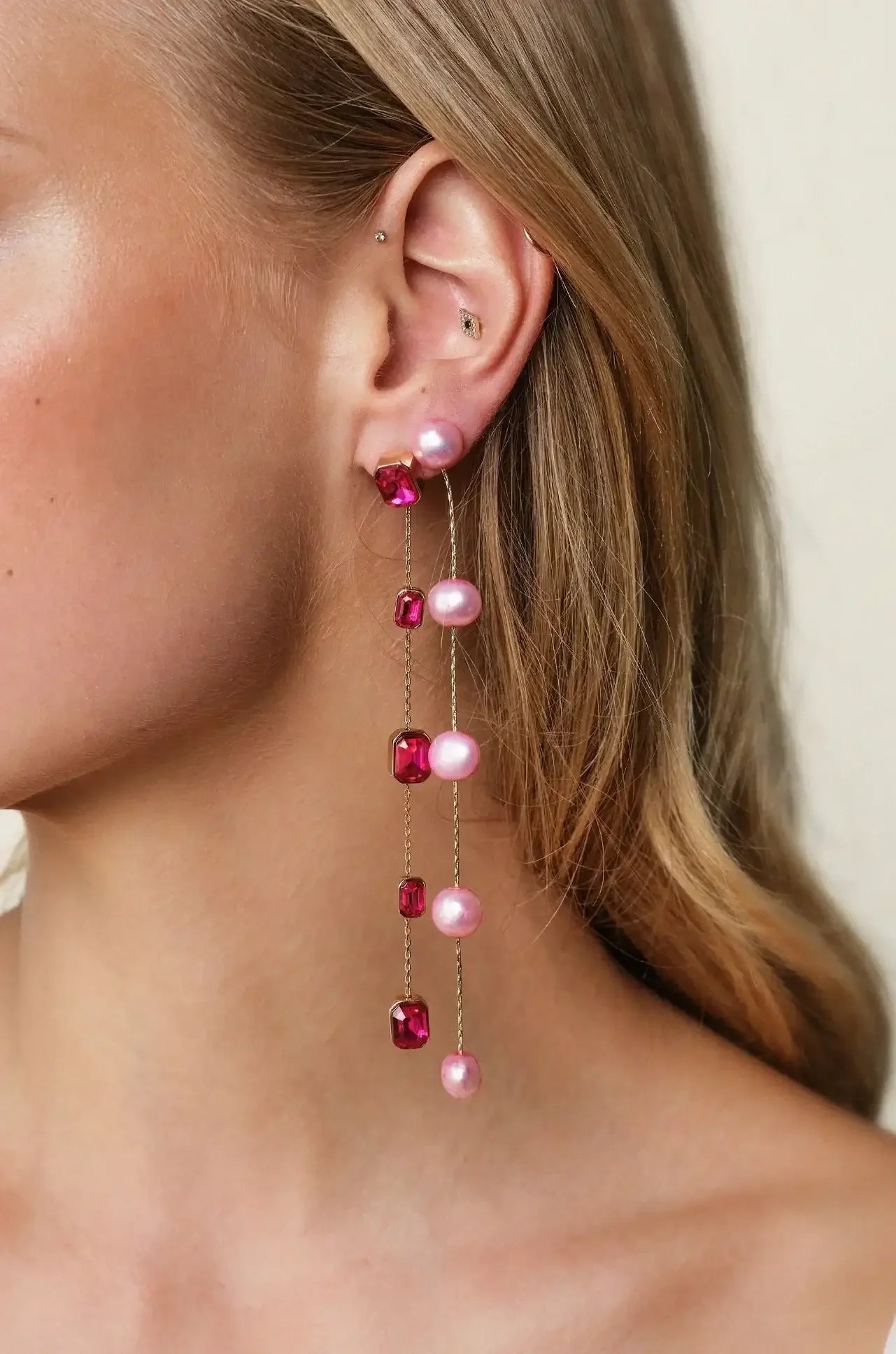 A dangling earring featuring pink pearls and red gemstones adorns a woman's ear, accentuating her long, flowing hair and smooth skin against a neutral background.