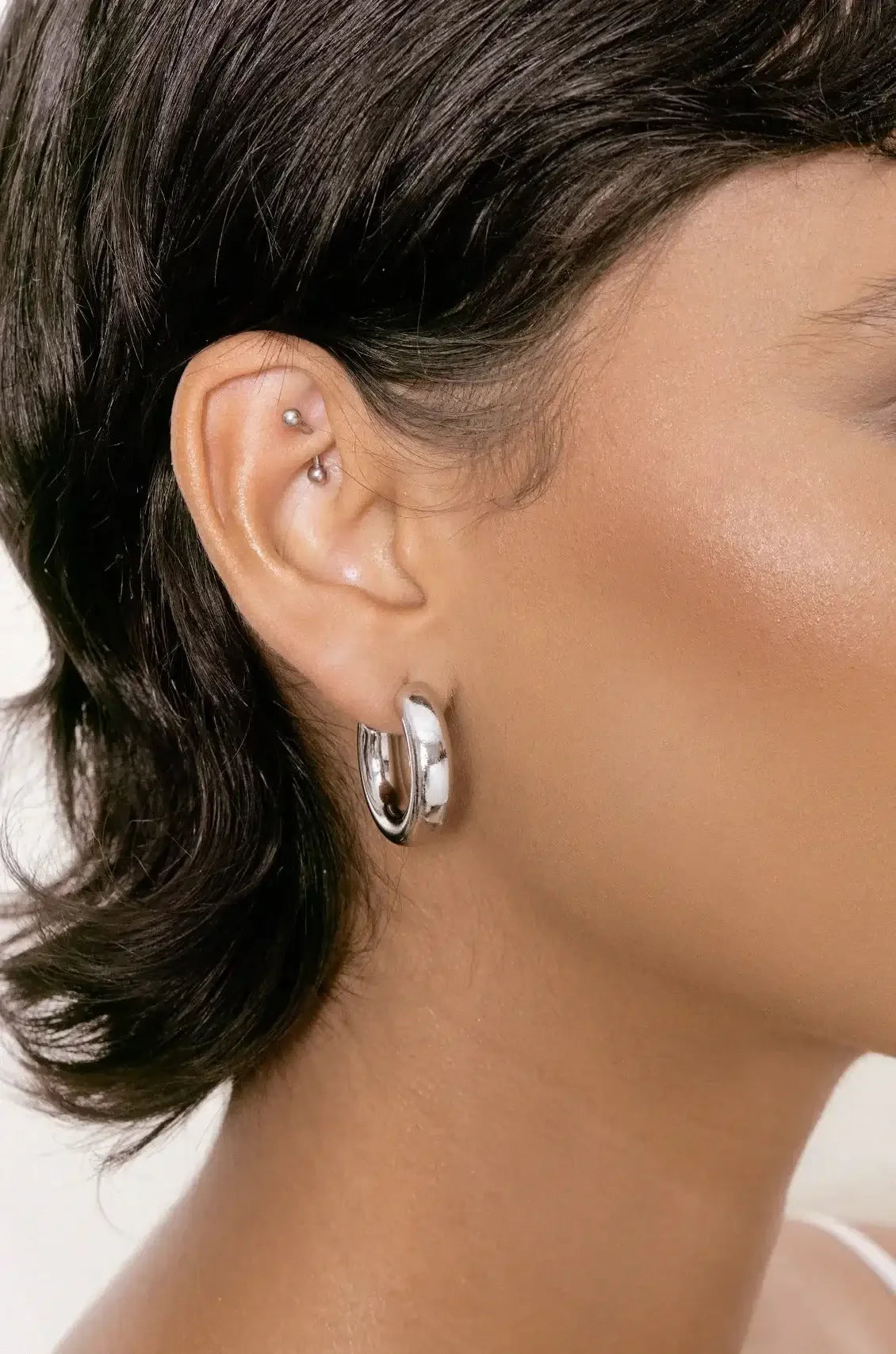 A close-up of a woman's ear, featuring a large silver hoop earring. Her short, dark hair frames her face, highlighting her skin's smoothness against a neutral background.