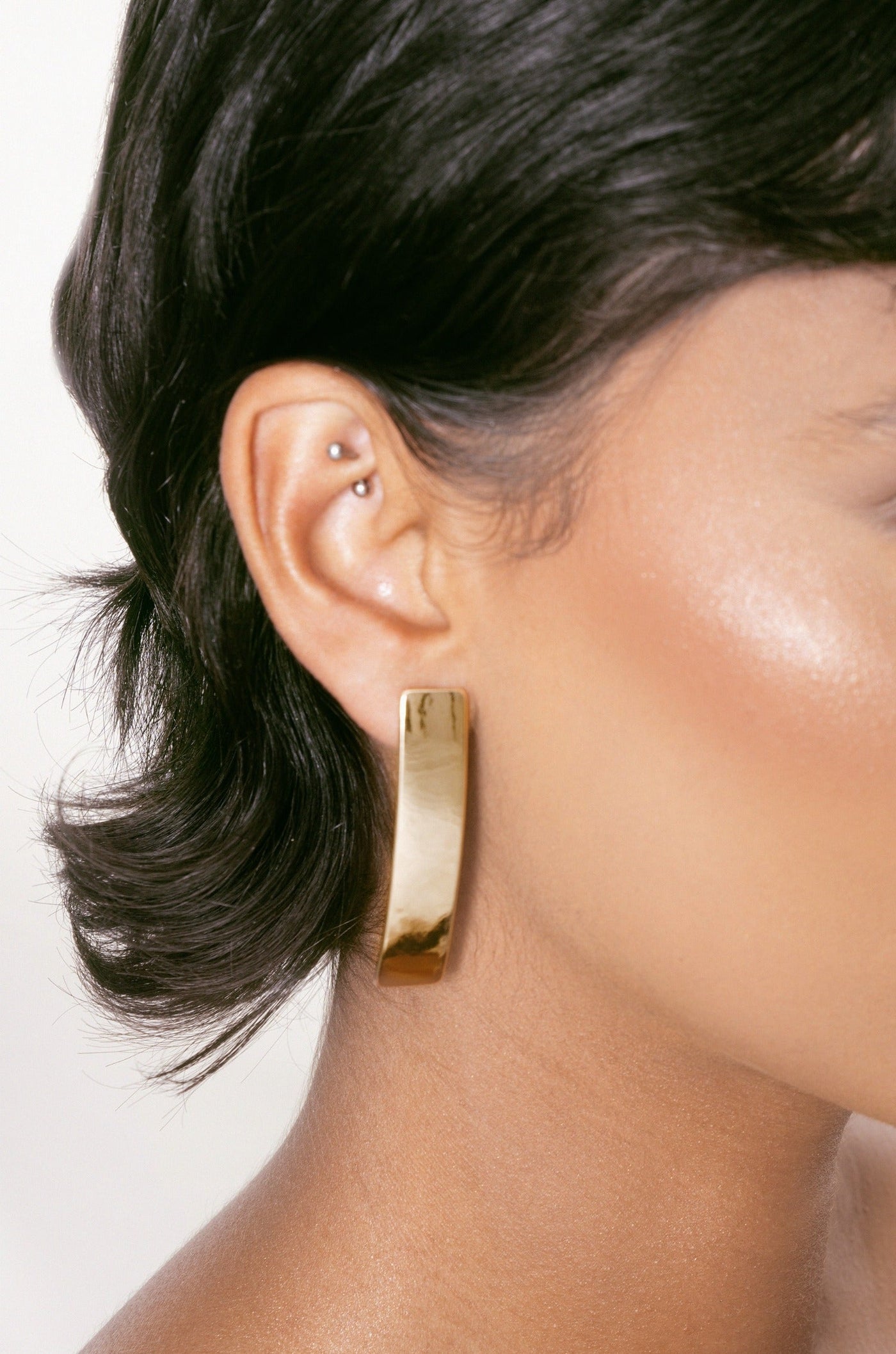 A large, shiny gold earring hangs from a pierced ear, while the person's smooth, illuminated skin and stylish short hair enhance the overall appearance against a plain light background.