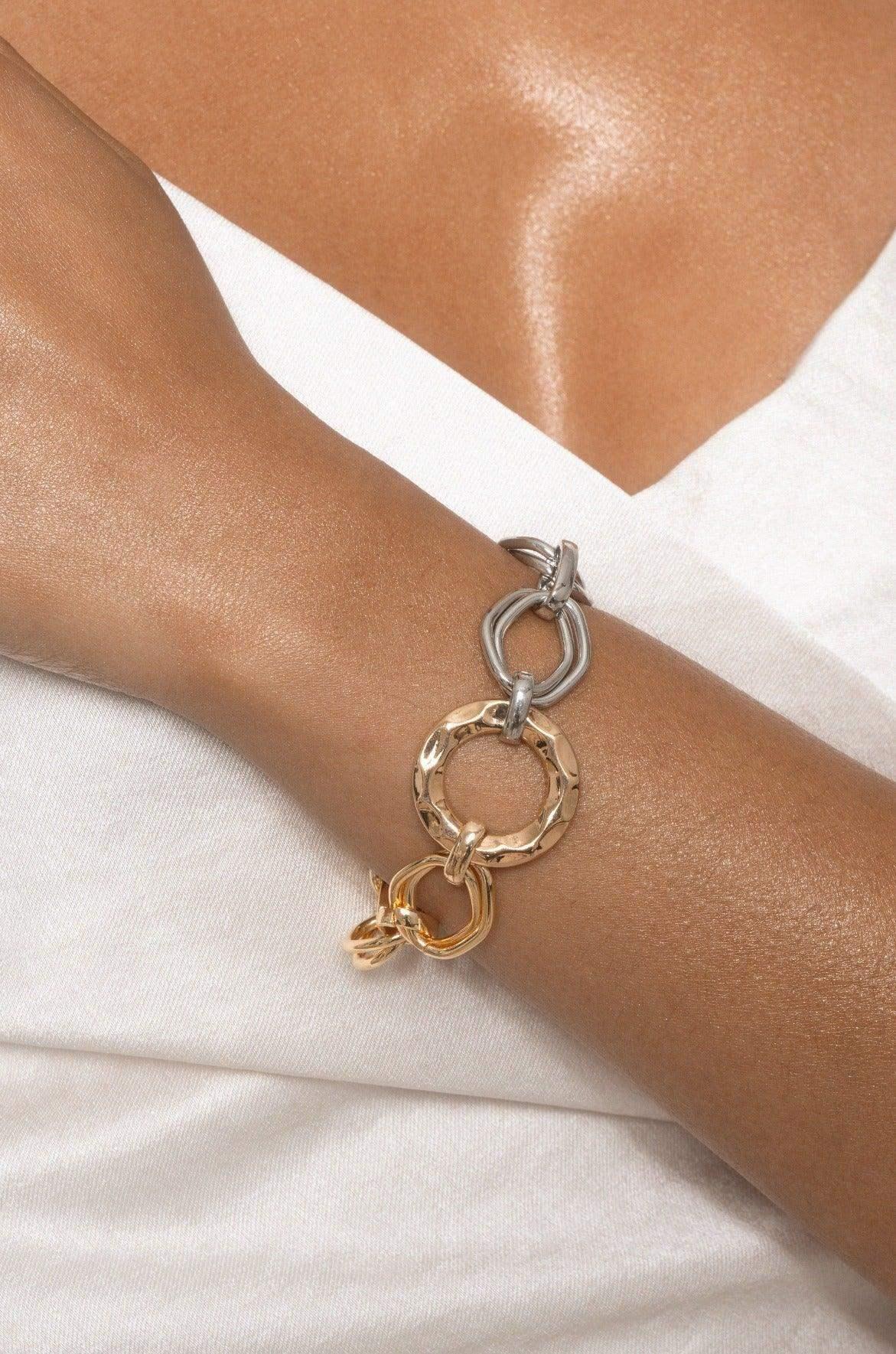 A close-up of a wrist showcases a fashionable bracelet made of intertwined metallic rings—silver, gold, and a textured circle—against smooth, tanned skin, with a white top in the background.