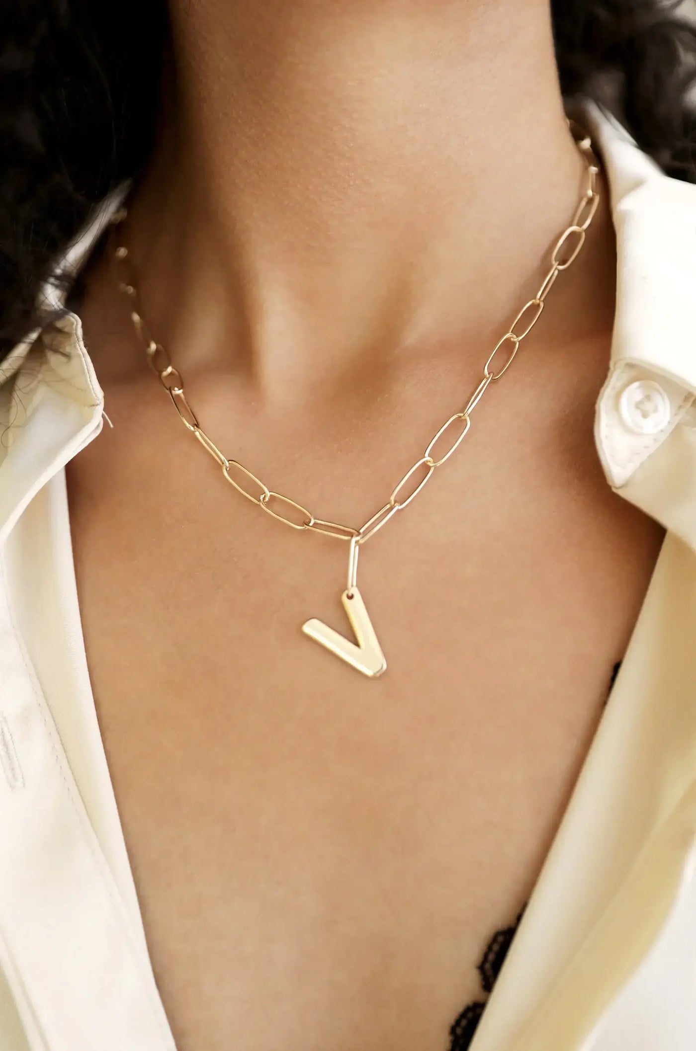 A gold chain necklace with a “V†pendant hangs around the neck, complementing a collared white shirt. The skin is smooth and the shirt is slightly unbuttoned, revealing lace underneath.
