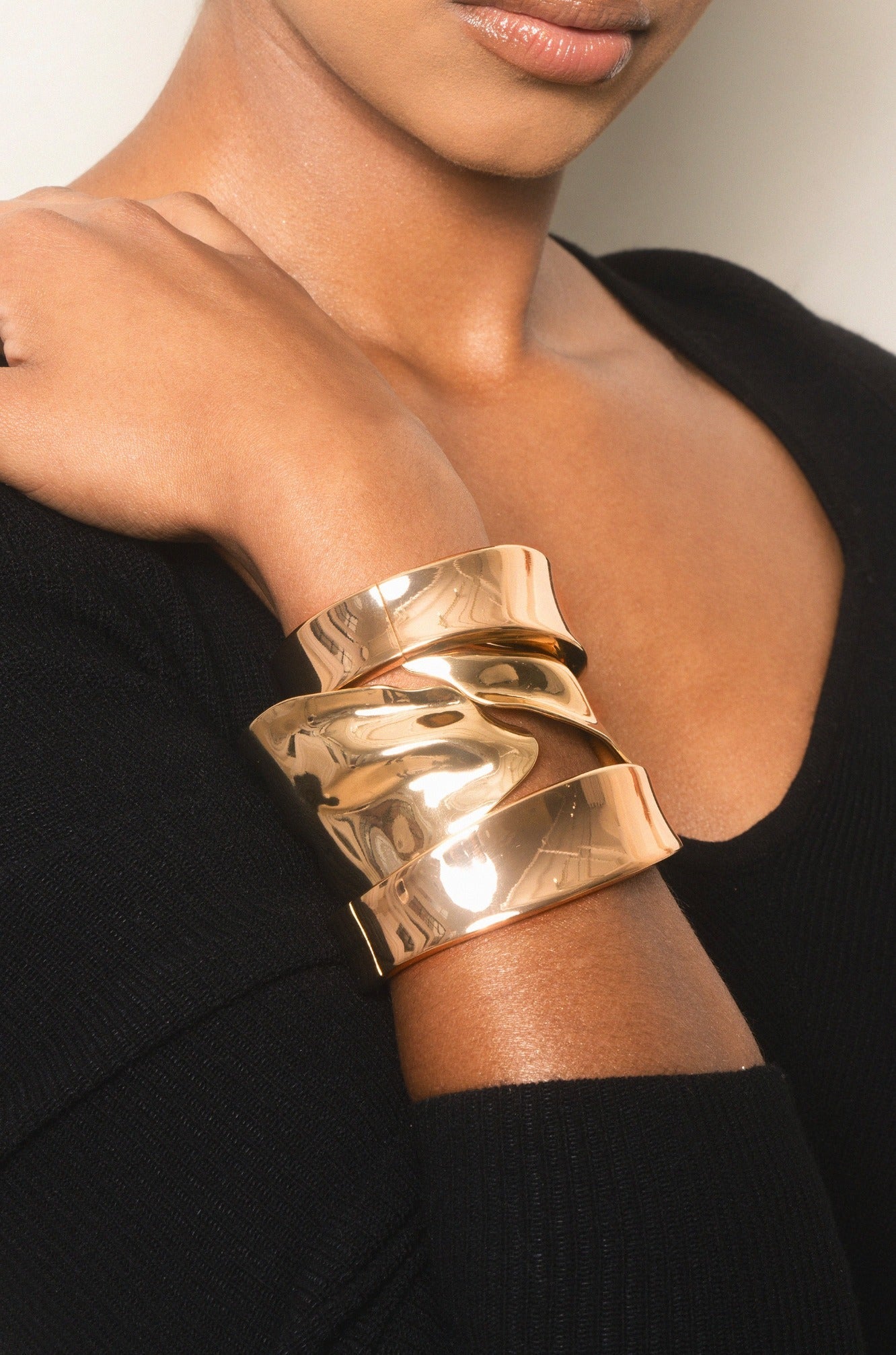 A gold-toned cuff bracelet adorned with wavy layers is worn on a person's wrist. The individual is partially visible, wearing a black sweater. The background appears neutral and unobtrusive.