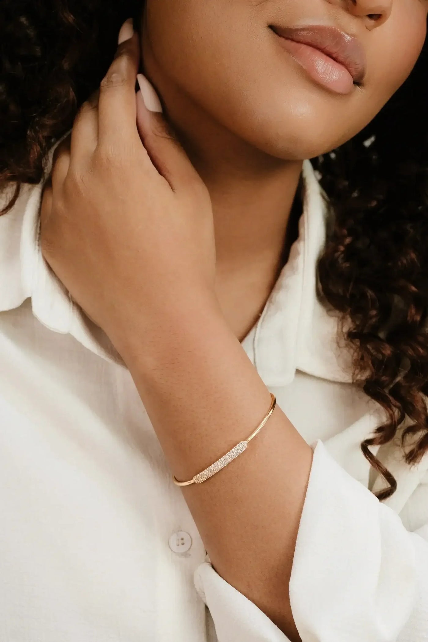 A hand gently touches the neck of a person wearing a light-colored shirt. A gold bracelet with a textured detail adorns the wrist, highlighting the accessory against the skin.