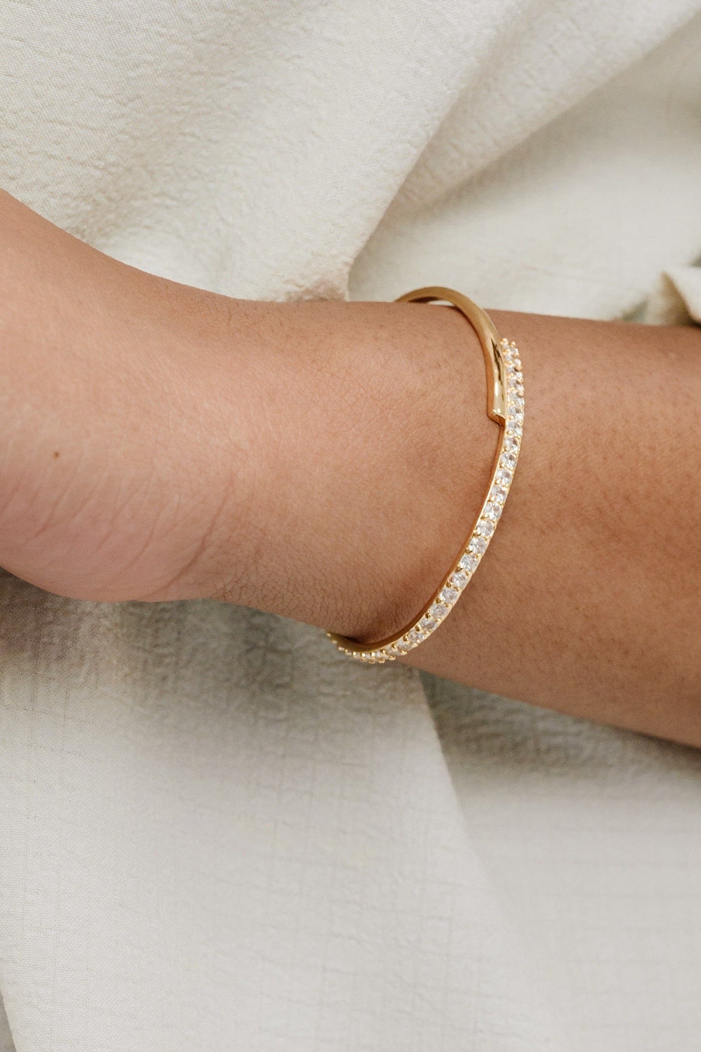 A gold bracelet adorned with small sparkly stones is worn on a wrist. The skin is a warm tone, set against a light, textured fabric background.