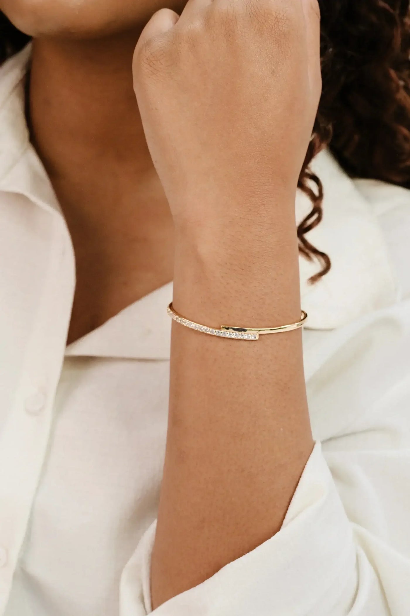 A gold bracelet with a sparkling crystal detail adorns a wrist, while the hand is raised slightly, showcasing the jewelry against a soft, light-colored shirt in a neutral setting.
